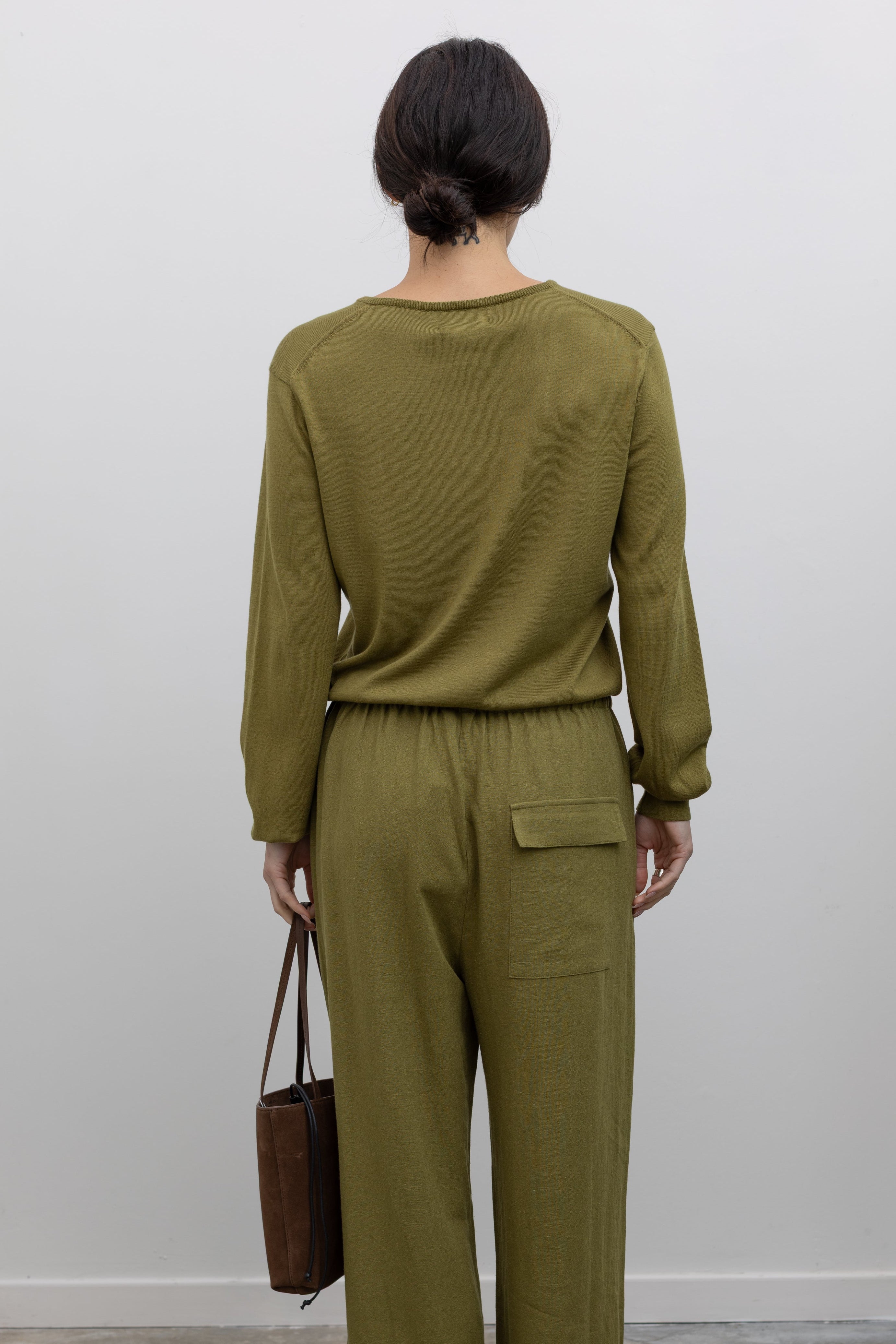 Full length back view of a woman posing wearing elevated capsule wardrobe essentials in the Mijeong Park Soft Green Cotton Blend V-Neck Knit Long Sleeve Top paired with Mijeong Park Green Linen Blend Wide Leg Cropped Pants featuring a comfortable drawstring waist, front and back patch pockets, and a cropped length suitable for a minimalist, versatile wardrobe at an Alas Eius store. 