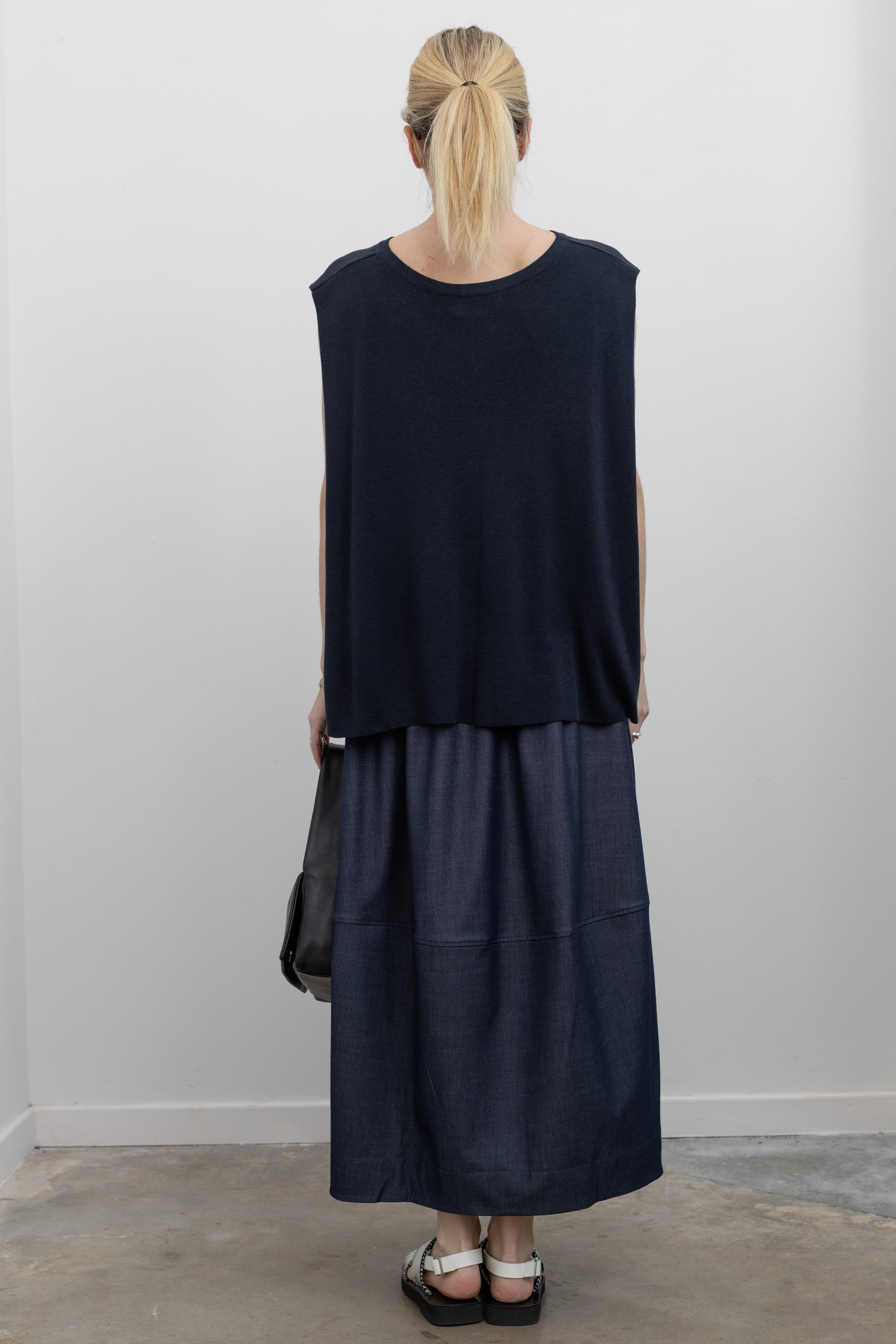 Full length back view of a woman standing wearing a modern and modest Mijeong Park brand navy blue sleeveless top with a fluid draped back and a curved front hem paired with a dark wash denim midi skirt at an Alas Eius store. 