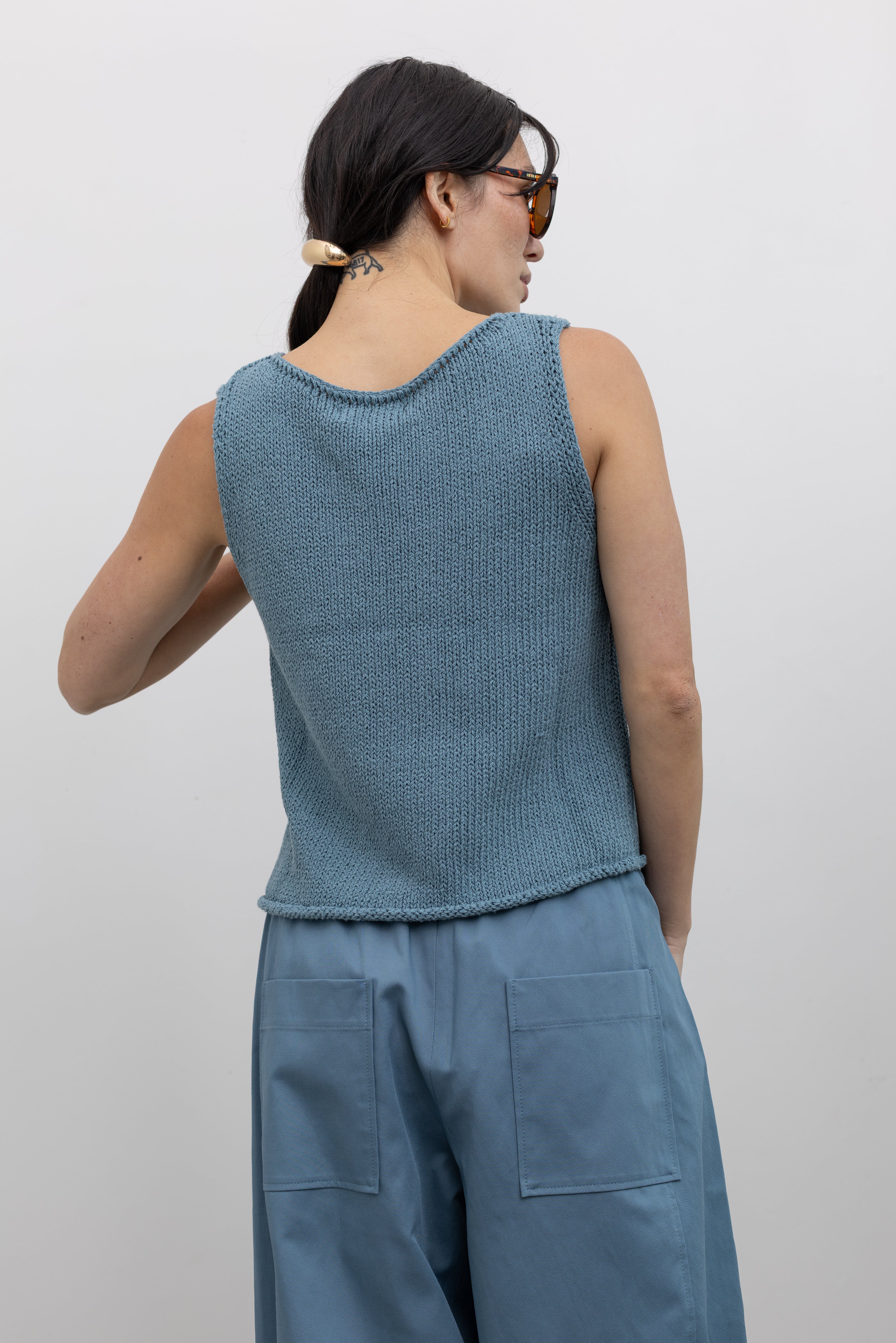 Close-up back view of a woman wearing elevated and modern spring fashion in a Mijeong Park light blue boat neck knit sweater tank top featuring rolled edges around the neckline and hem at an Alas Eius store. 