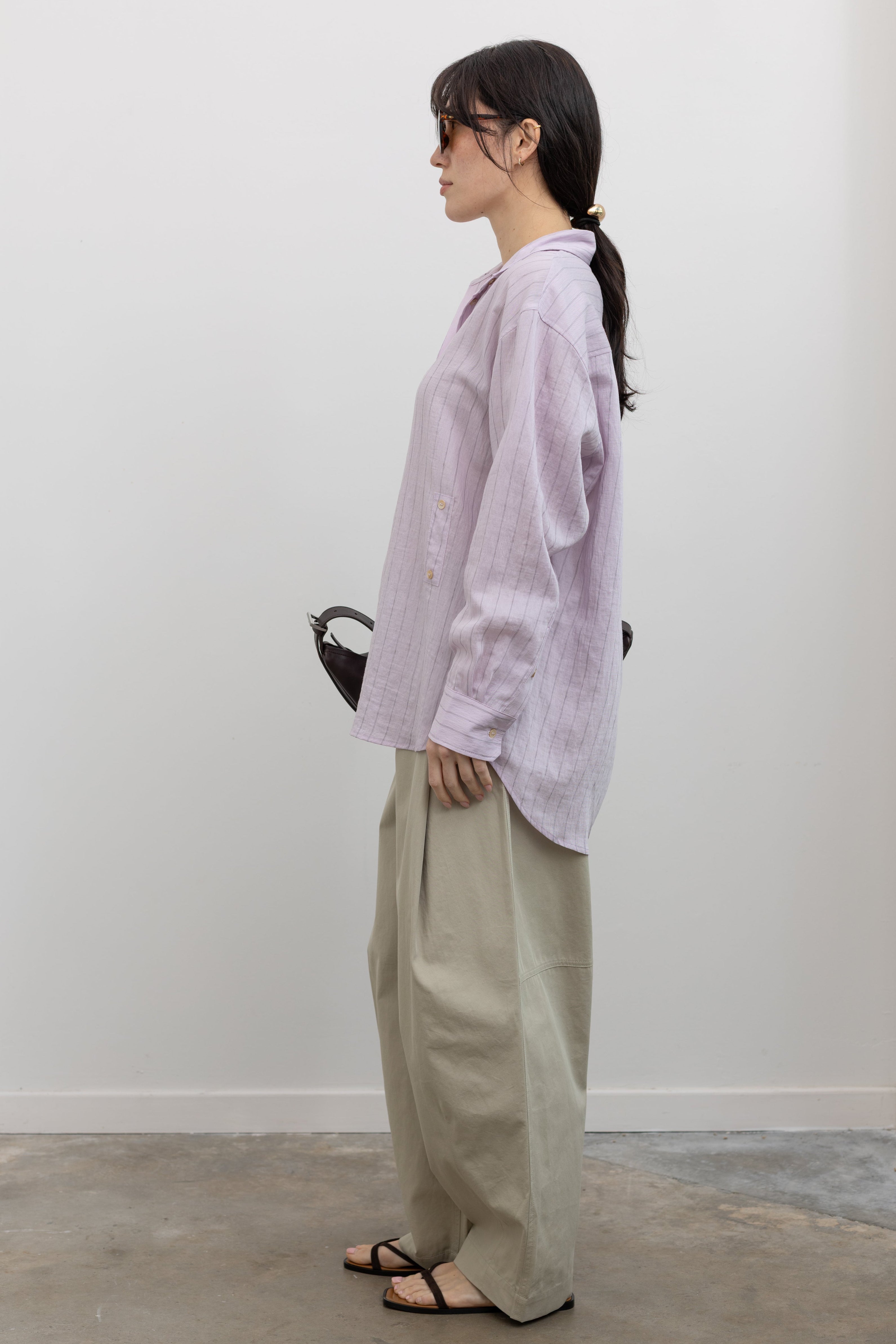 Full length side view of a woman wearing modern minimalist spring fashion in a Mijeong Park oversized soft pink pinstripe button down shirt featuring a classic collar, long sleeves, and an off center button down front paired with an elevated classic khaki baggy curved leg pant with strappy sandals at an Alas Eius store. 