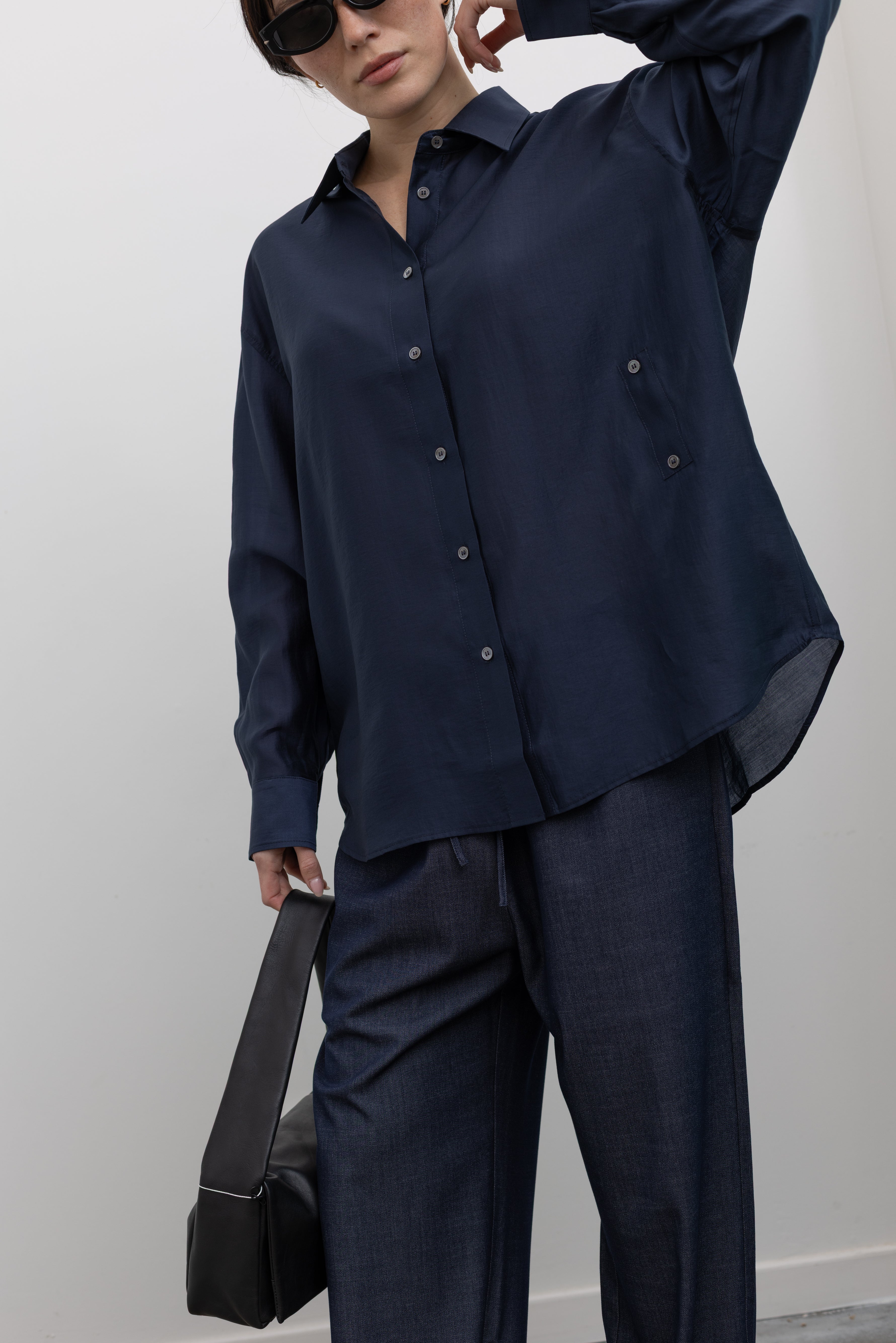 Close-up front view of a woman wearing a modern minimalist staple in a Mijeong Park navy blue semi-sheer oversized shirt featuring a flattering off-center button front, long sleeves, and a classic sharp collar paired with Mijeong Park lightweight dark wash denim drawstring pants, Salomon slide sneakers, and a leather St. Agni bag at an Alas Eius store. 