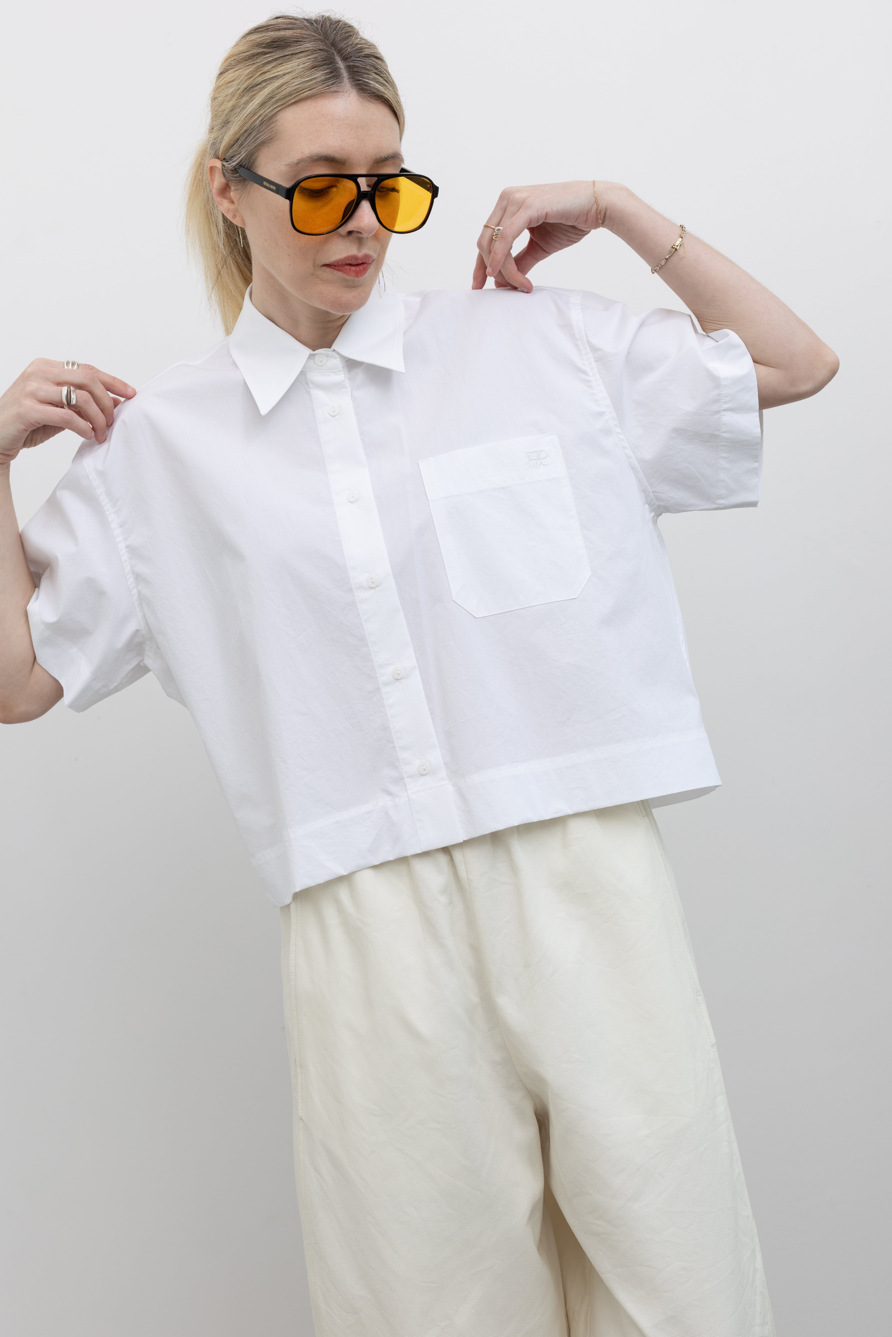 Front view of a woman posing with sunglasses wearing modern minimalist spring fashion in a Mijeong Park 100% cotton white oversized collared button down shirt featuring a chest patch pocket, a cropped hem, and short sleeves paired with an elevated classic wardrobe staple in Mijeong Park cream baggy curved leg trousers at an Alas Eius store.