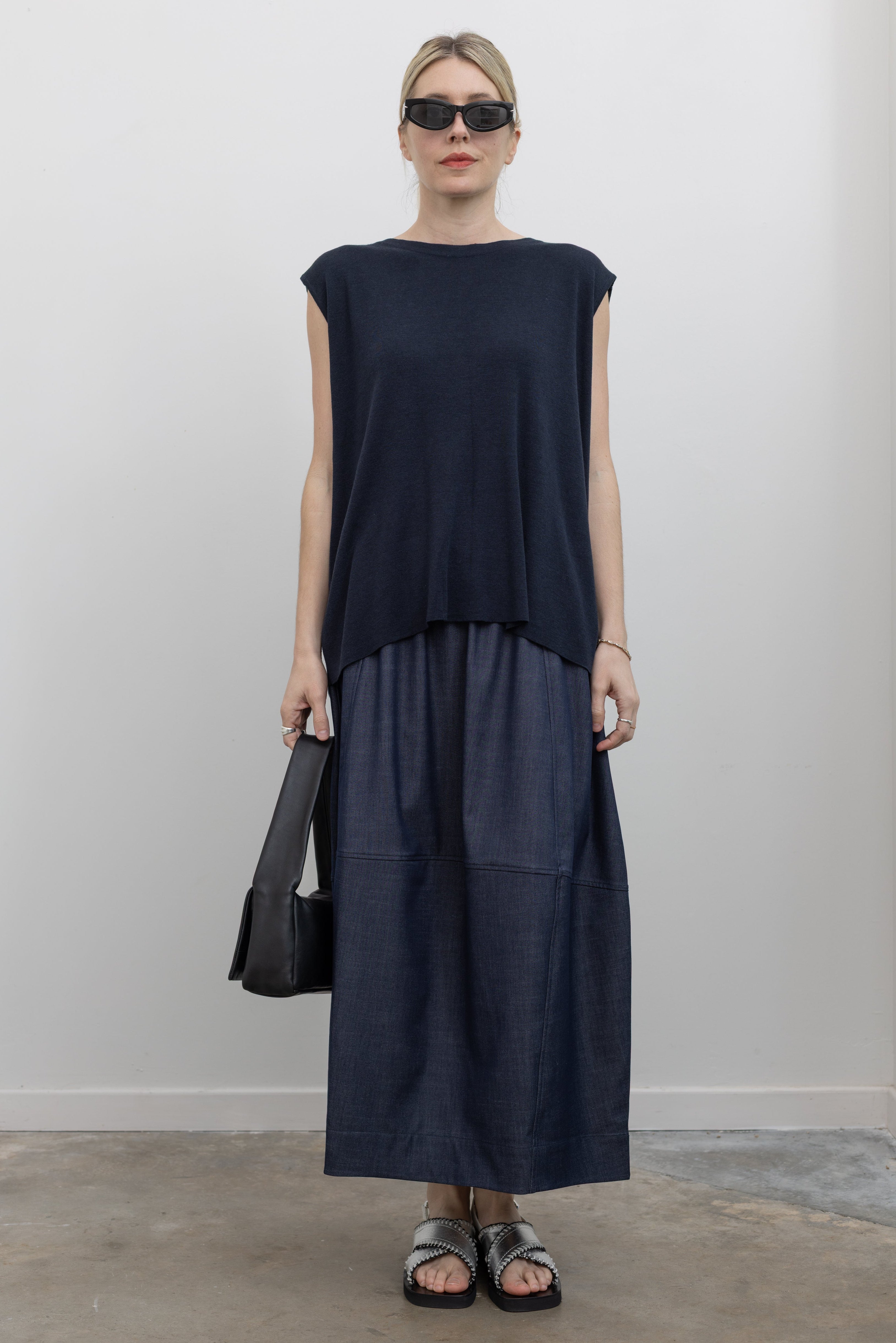 Full length front view of a woman standing wearing modest spring fashion in a Mijeong Park navy drape back knit top with a dark wash denim blue voluminous balloon midi-to-maxi silhouette skirt featuring an elastic waistband paired with silver metallic slip-on sandals and a sleek St. Agni leather shoulder bag at an Alas Eius store. 