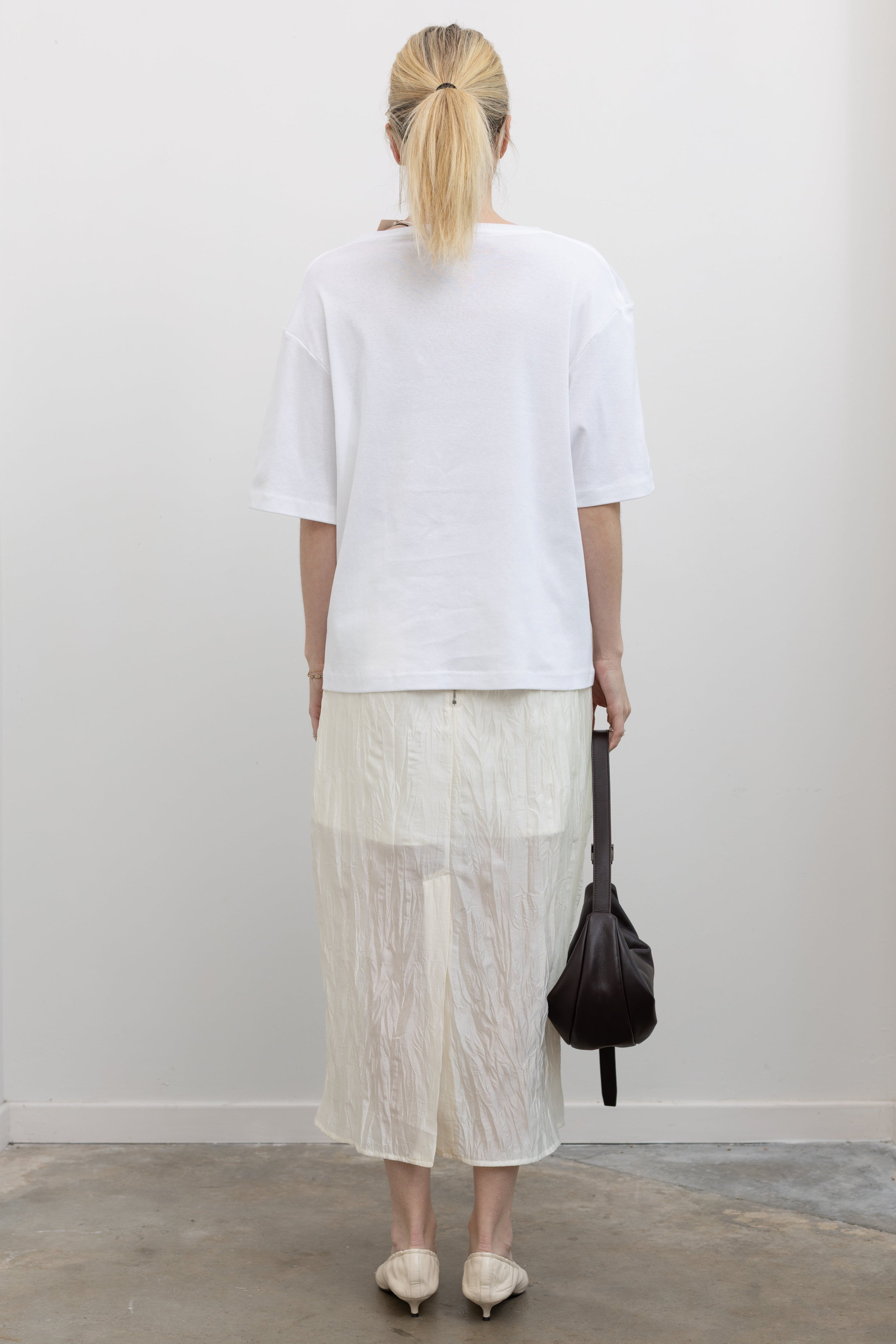 Full length back view of a woman standing wearing chic monochromatic spring fashion in a Mijeong Park white lightweight, semi-sheer crinkle fabric lined midi skirt paired with a Mijeong Park white oversized t-shirt at an Alas Eius store. 
