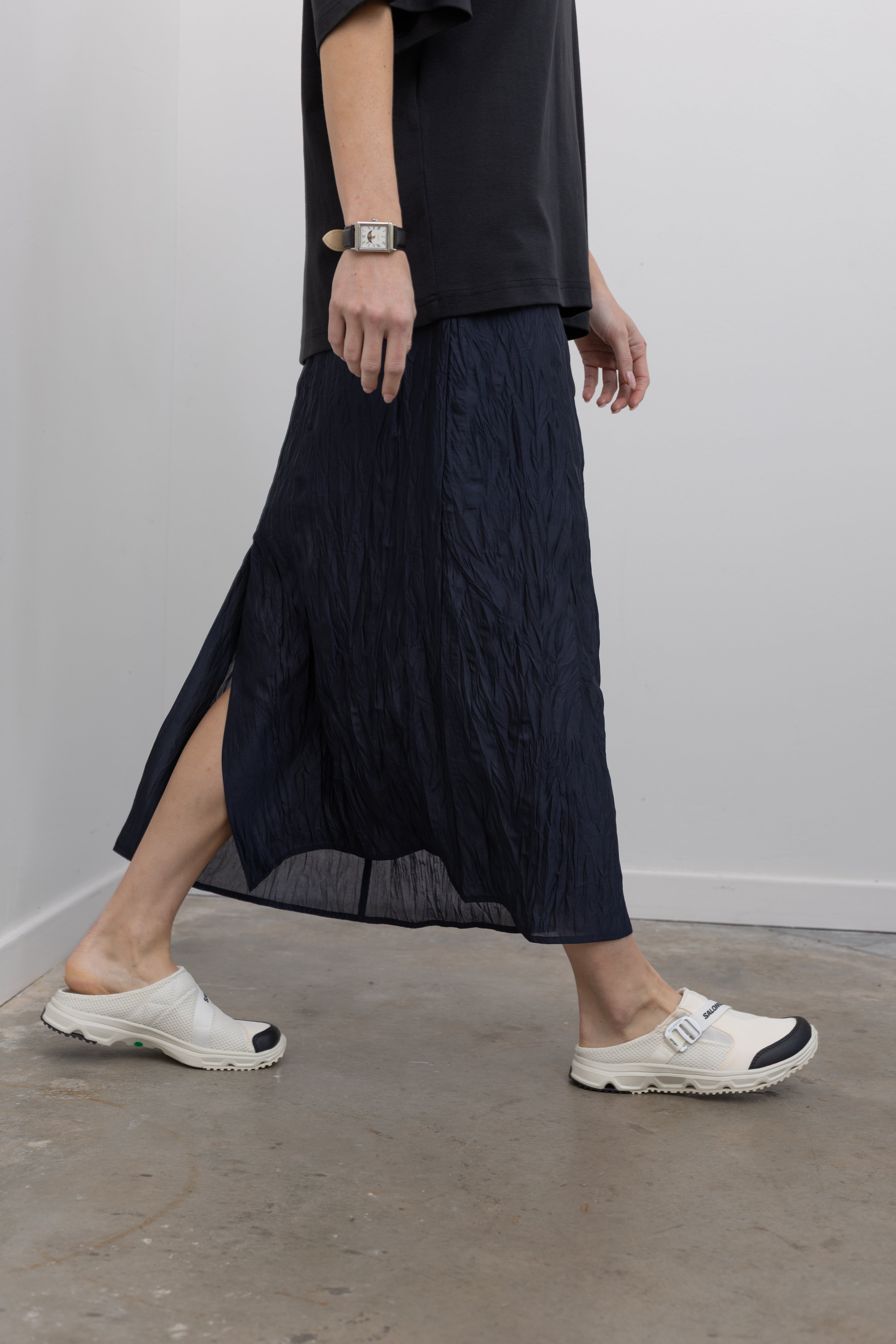 Close-up side view of a woman in motion wearing chic and versatile spring fashion in a Mijeong Park navy blue lightweight, semi-sheer crinkled fabric lined midi skirt paired with a Mijeong Park black oversized t-shirt and Salomon sneakers at an Alas Eius store. 