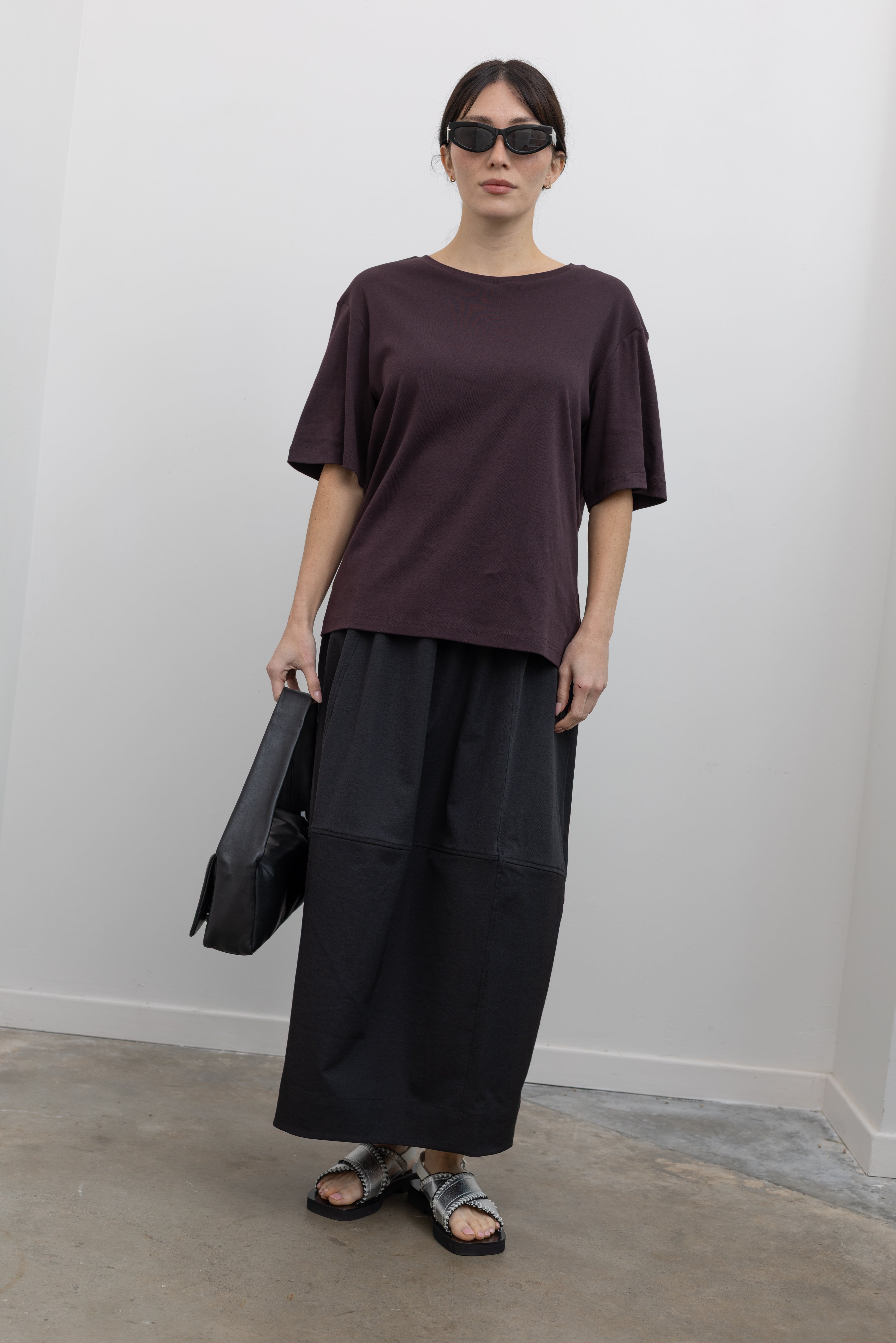 Full length front view of a woman standing wearing modest spring fashion in a Mijeong Park dark brown oversized t-shirt with a black voluminous balloon midi-to-maxi silhouette skirt featuring an elastic waistband paired with silver metallic slip-on sandals and a sleek St. Agni black leather shoulder bag at an Alas Eius store. 