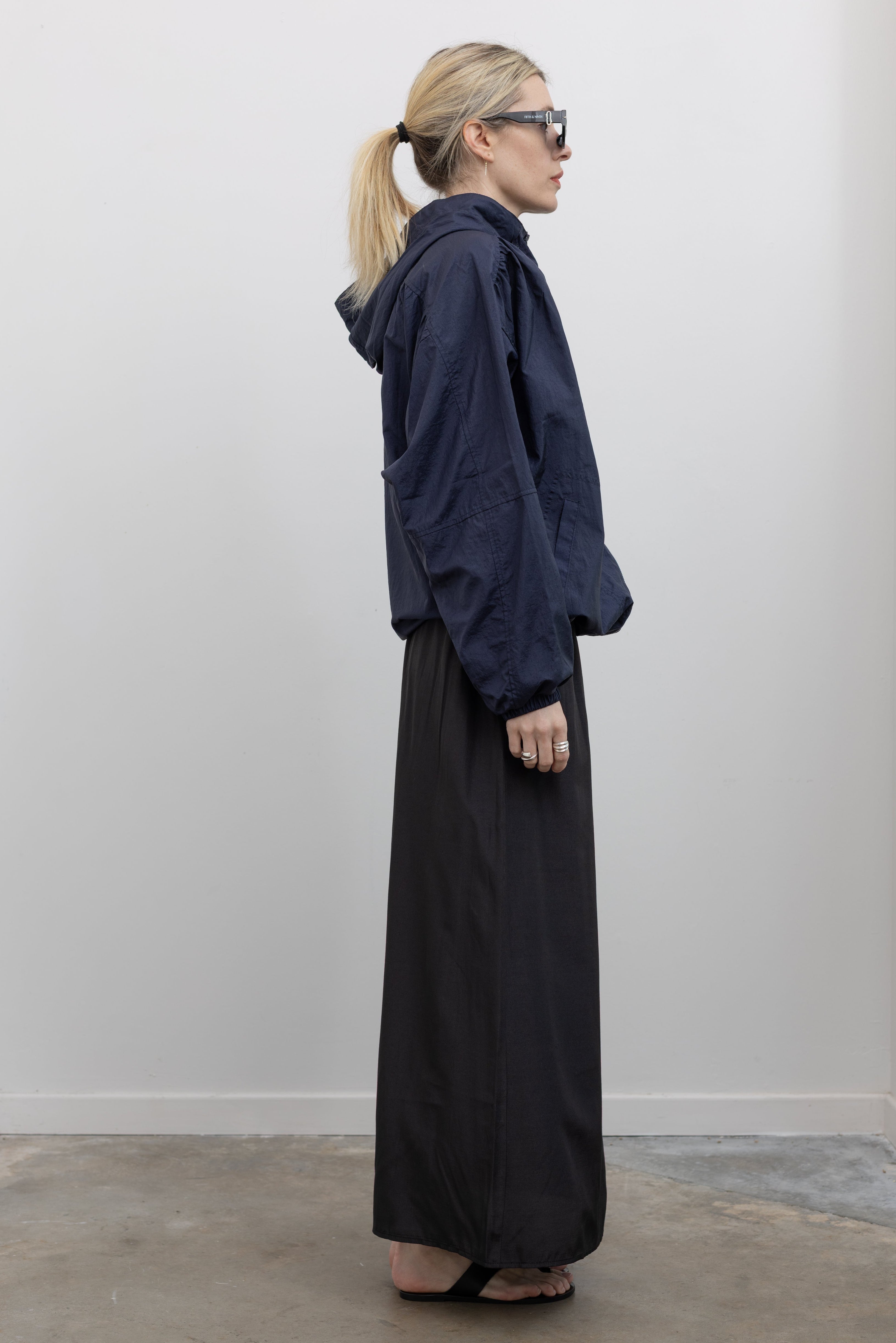 Full length side view of a woman wearing an elevated outerwear staple in a  Mijeong Park navy blue nylon shirred anorak jacket featuring an oversized fit, front pockets, hood, and a zipper closure at an Alas Eius store. 