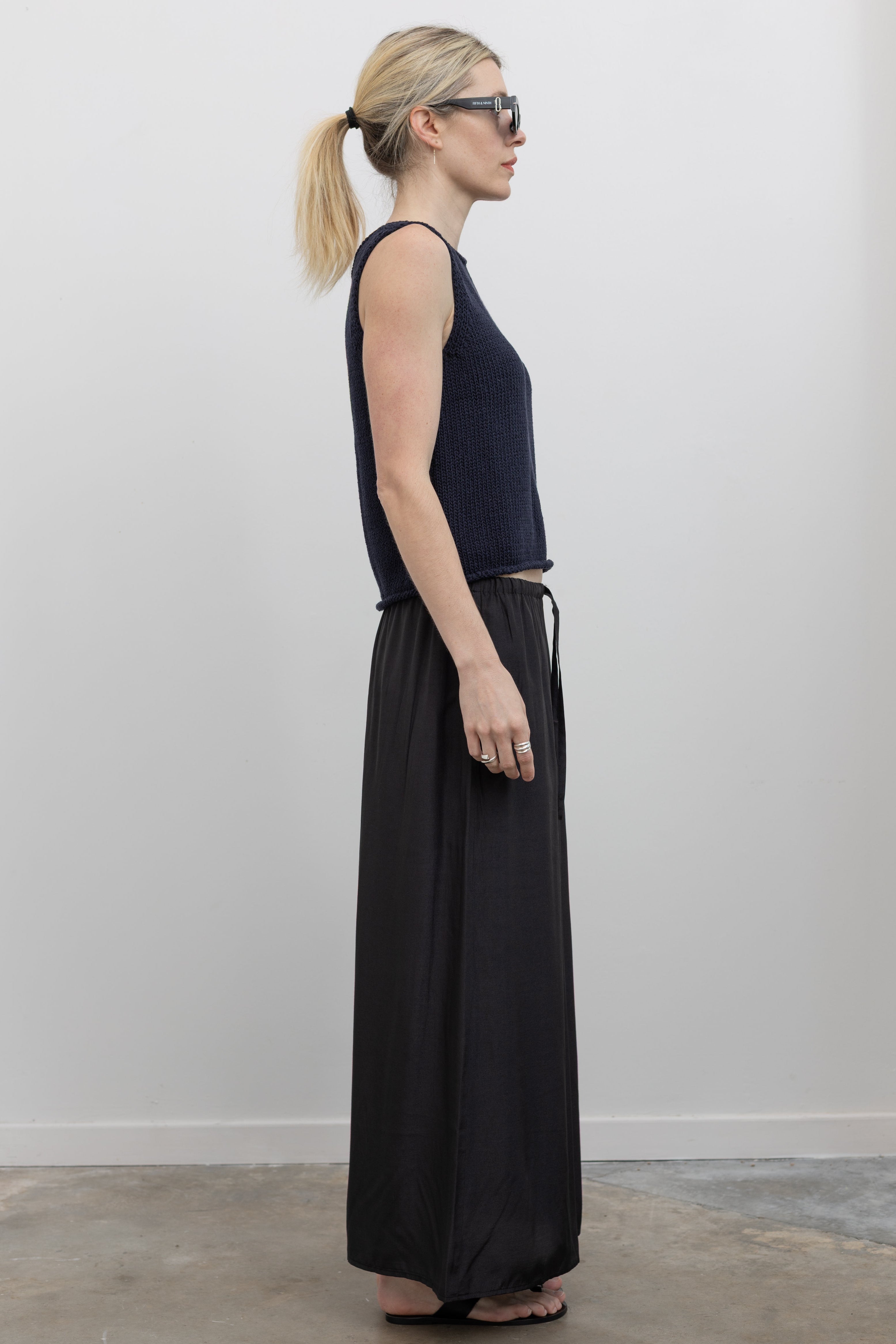 Full length side view of a woman standing wearing spring fashion in a Mijeong Park navy blue boat neck knit sweater tank top featuring rolled edges around the neckline and hem paired with a Mijeong Park black drawstring waist maxi skirt at an Alas Eius store. 