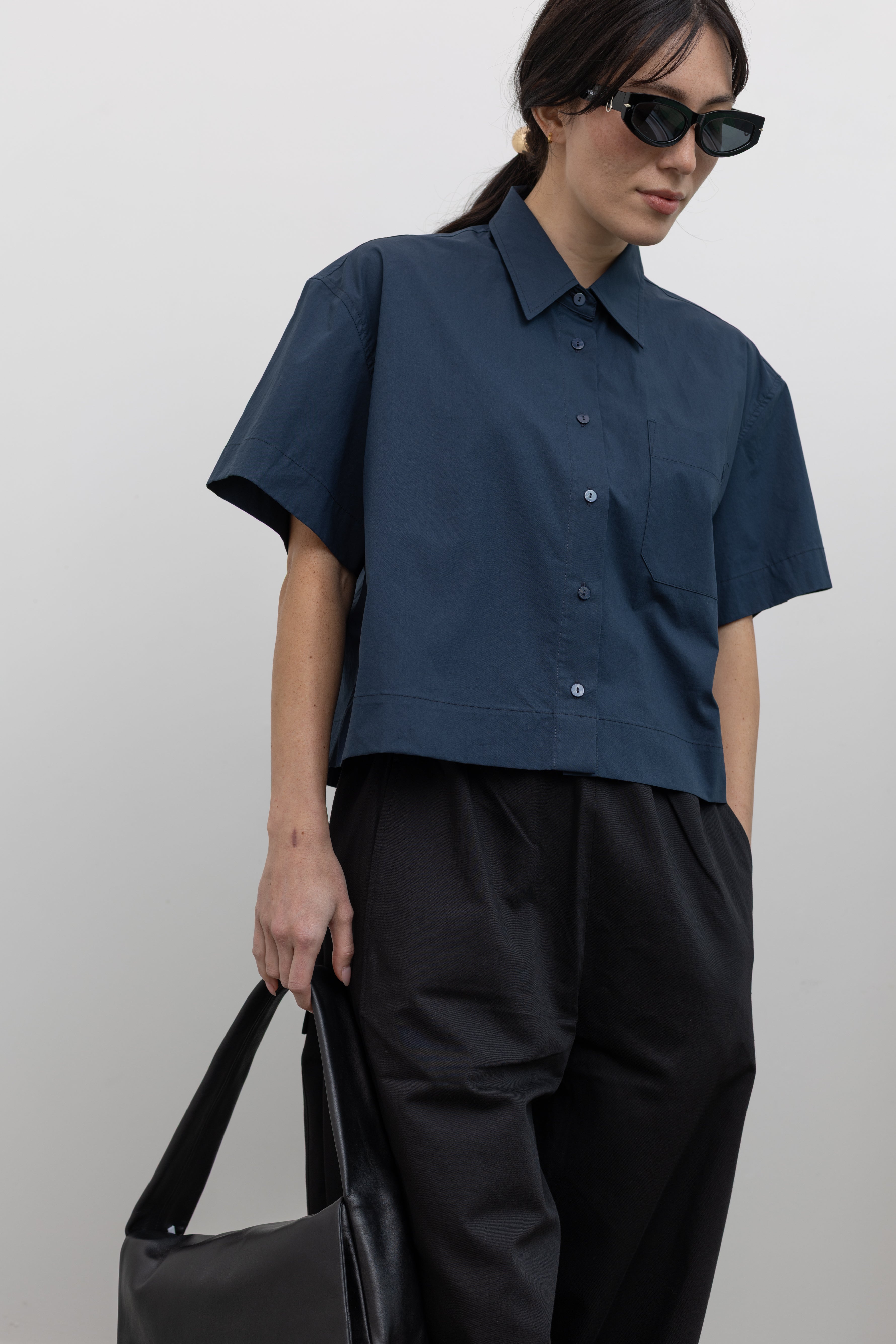 Front view of a woman posing with sunglasses wearing modern architectural spring fashion in a Mijeong Park 100% cotton navy blue oversized collared button down shirt featuring a chest patch pocket, a cropped hem, and short sleeves paired with an elevated classic wardrobe staple in Mijeong Park black baggy curved leg pants at an Alas Eius store.