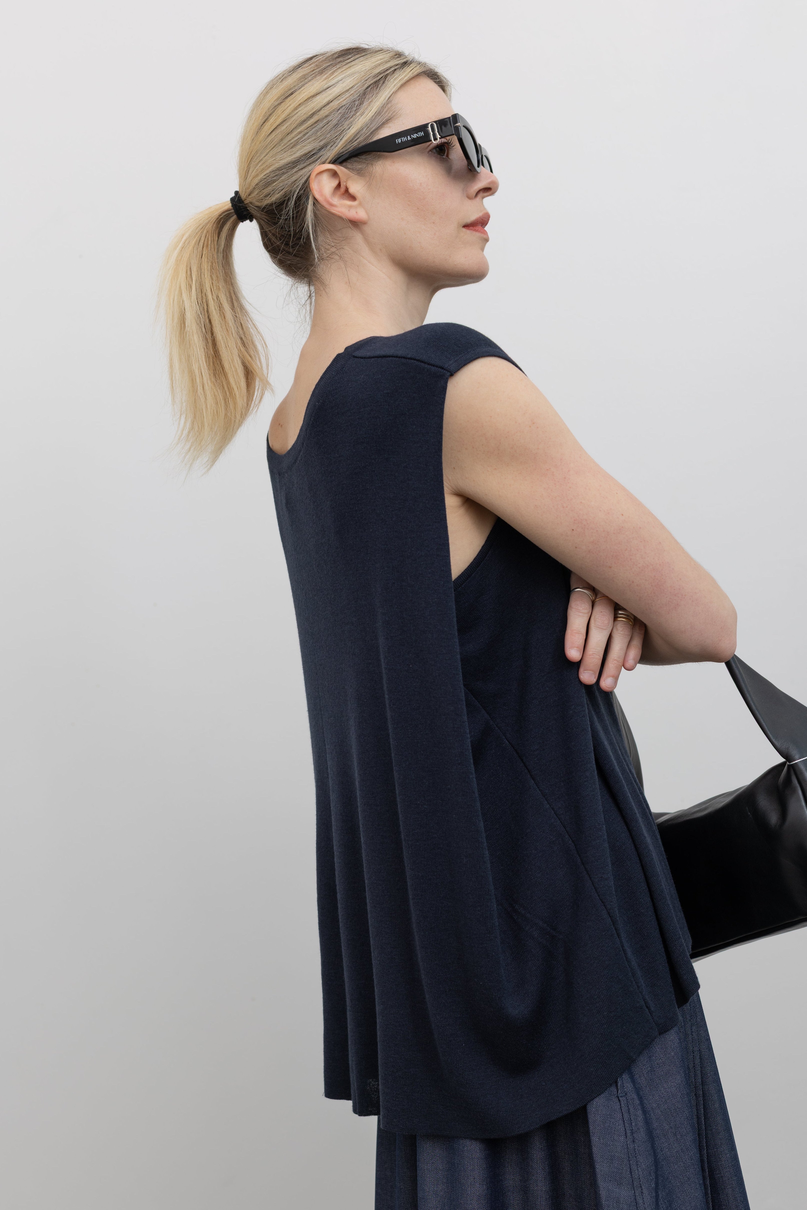 3/4 back view of a woman standing wearing flattering Mijeong Park brand navy blue sleeveless top with a fluid draped back at an Alas Eius store. 