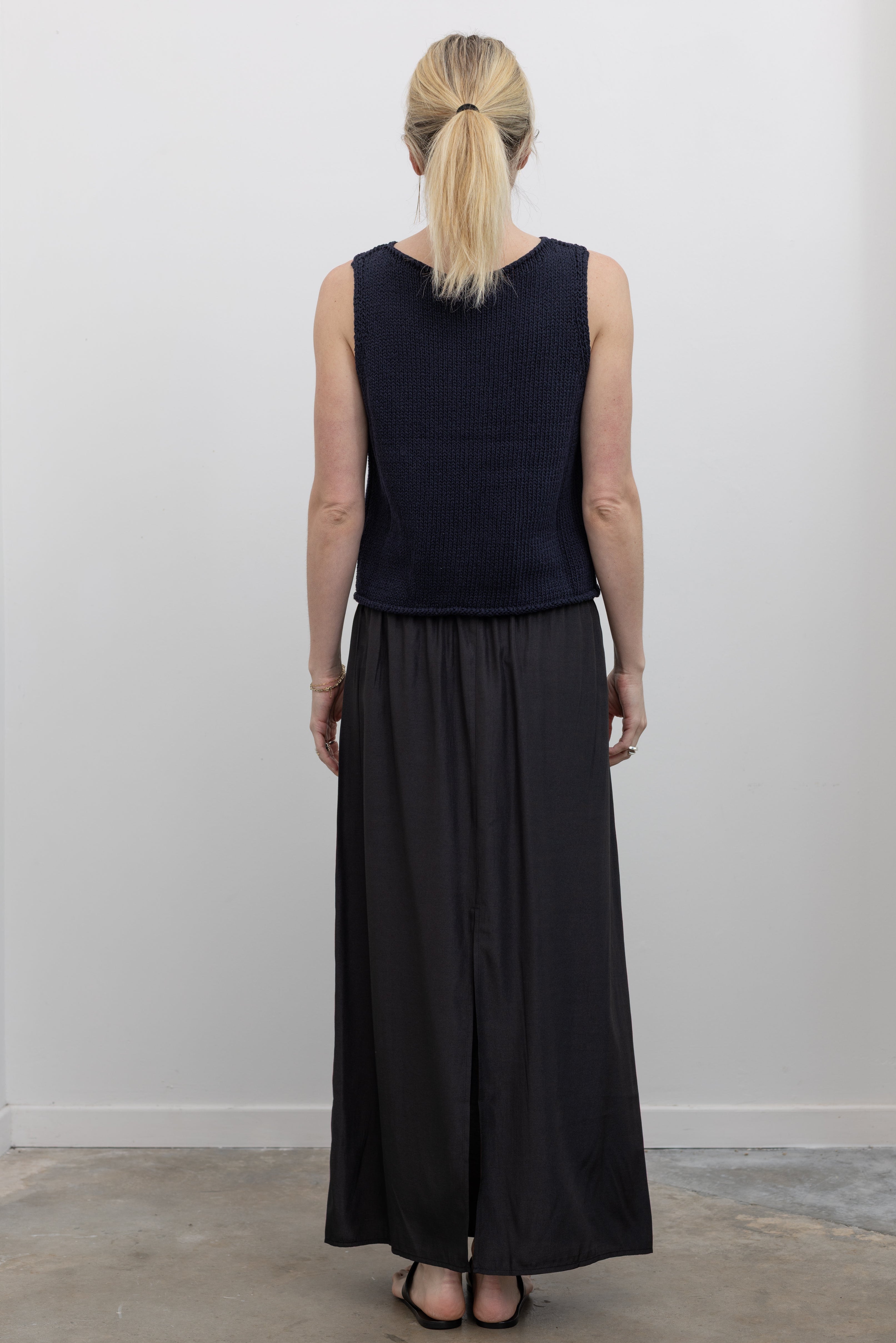 Full length back view of a woman standing wearing spring fashion in a Mijeong Park navy blue boat neck knit sweater tank top featuring rolled edges around the neckline and hem paired with a Mijeong Park black drawstring waist maxi skirt at an Alas Eius store. 