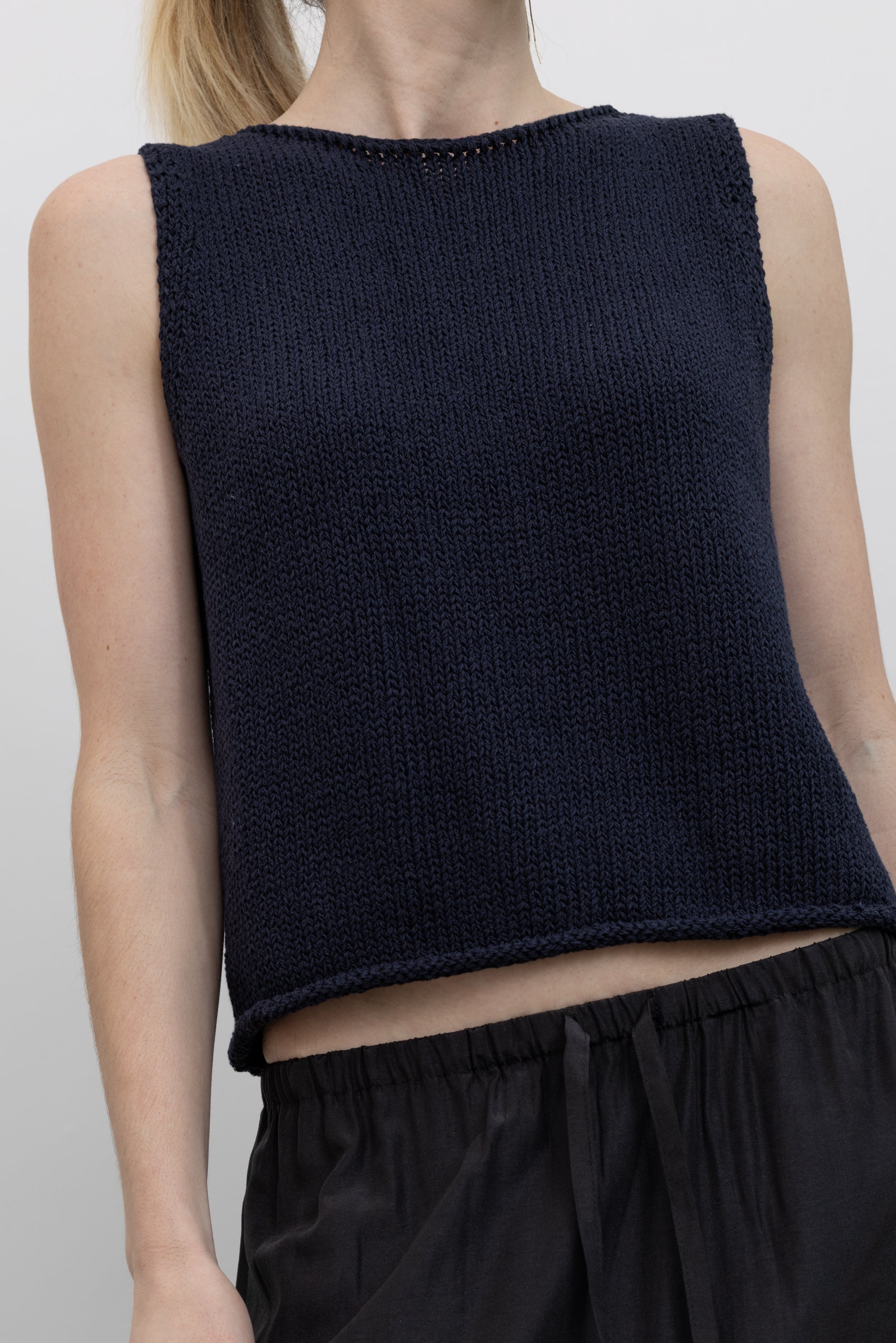 Close-up front view of a woman standing wearing spring fashion in a Mijeong Park navy blue boat neck knit sweater tank top featuring rolled edges around the neckline and hem paired with a Mijeong Park black drawstring waist maxi skirt at an Alas Eius store. 