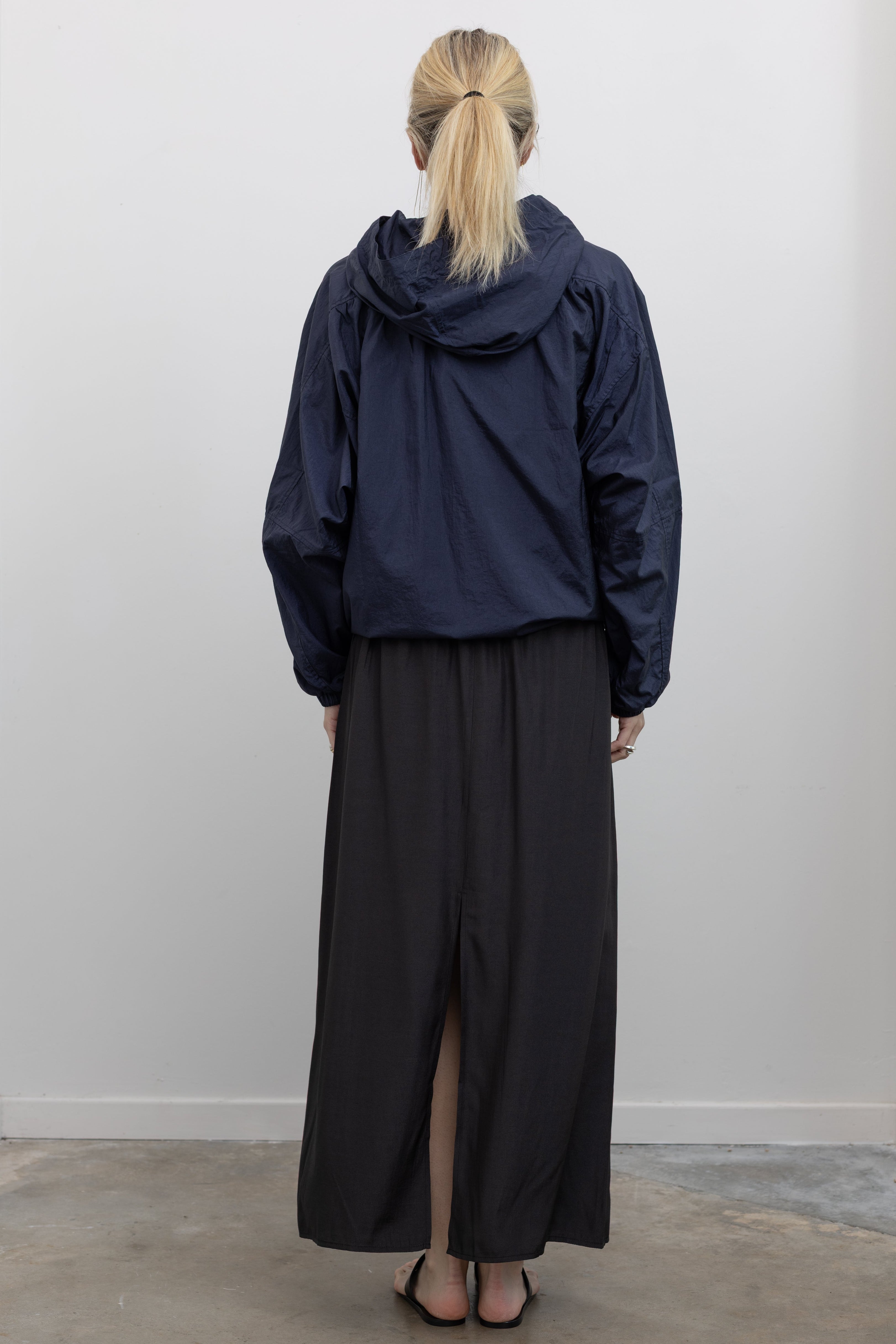 Full length back view of a woman wearing an elevated outerwear staple in a Mijeong Park navy blue nylon shirred anorak jacket featuring an oversized fit, front pockets, hood, and a zipper closure at an Alas Eius store. 