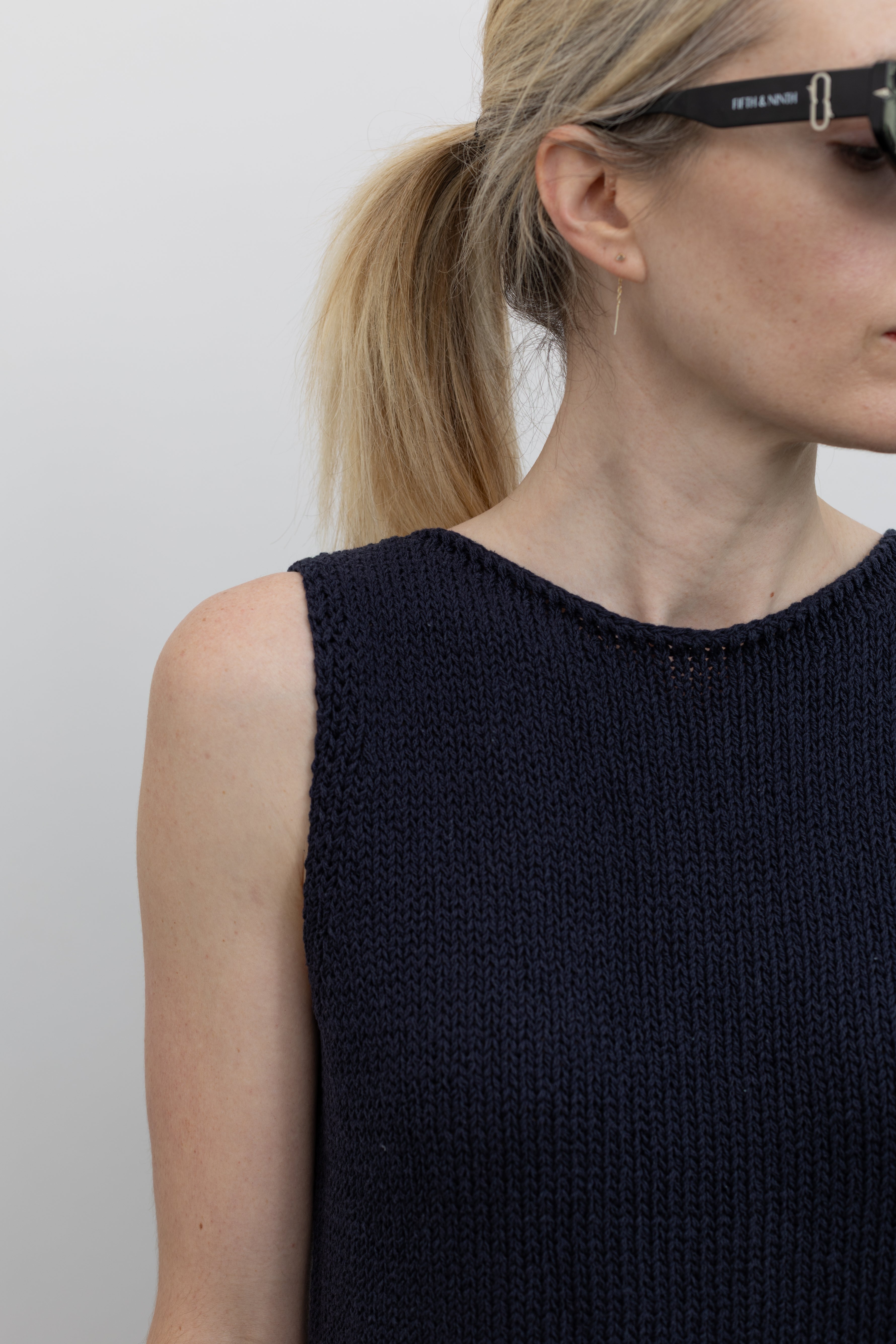 Close-up detailed front view of a woman standing wearing spring fashion in a Mijeong Park navy blue boat neck knit sweater tank top featuring rolled edges around the neckline and hem paired with a Mijeong Park black drawstring waist maxi skirt at an Alas Eius store. 