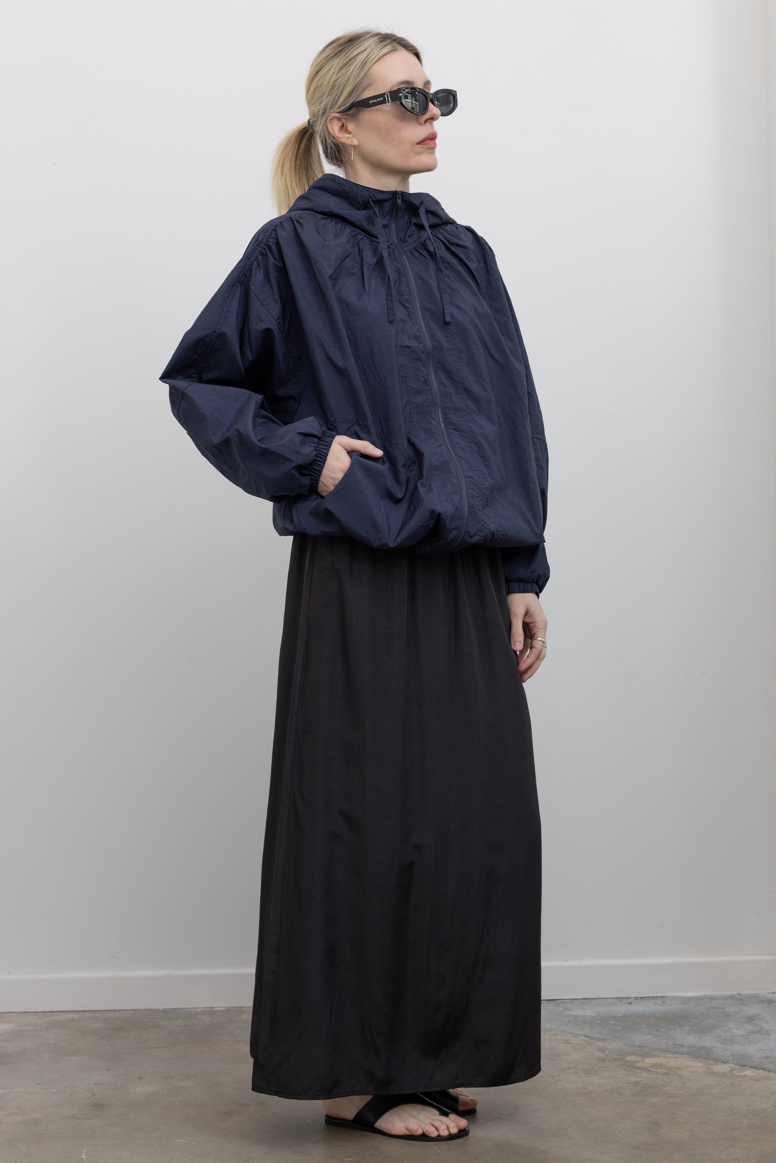 3/4 view of a woman posing with sunglasses wearing an versatile outerwear staple in a  Mijeong Park navy blue nylon shirred anorak jacket featuring an oversized fit, front pockets, hood, and a zipper closure at an Alas Eius store. 
