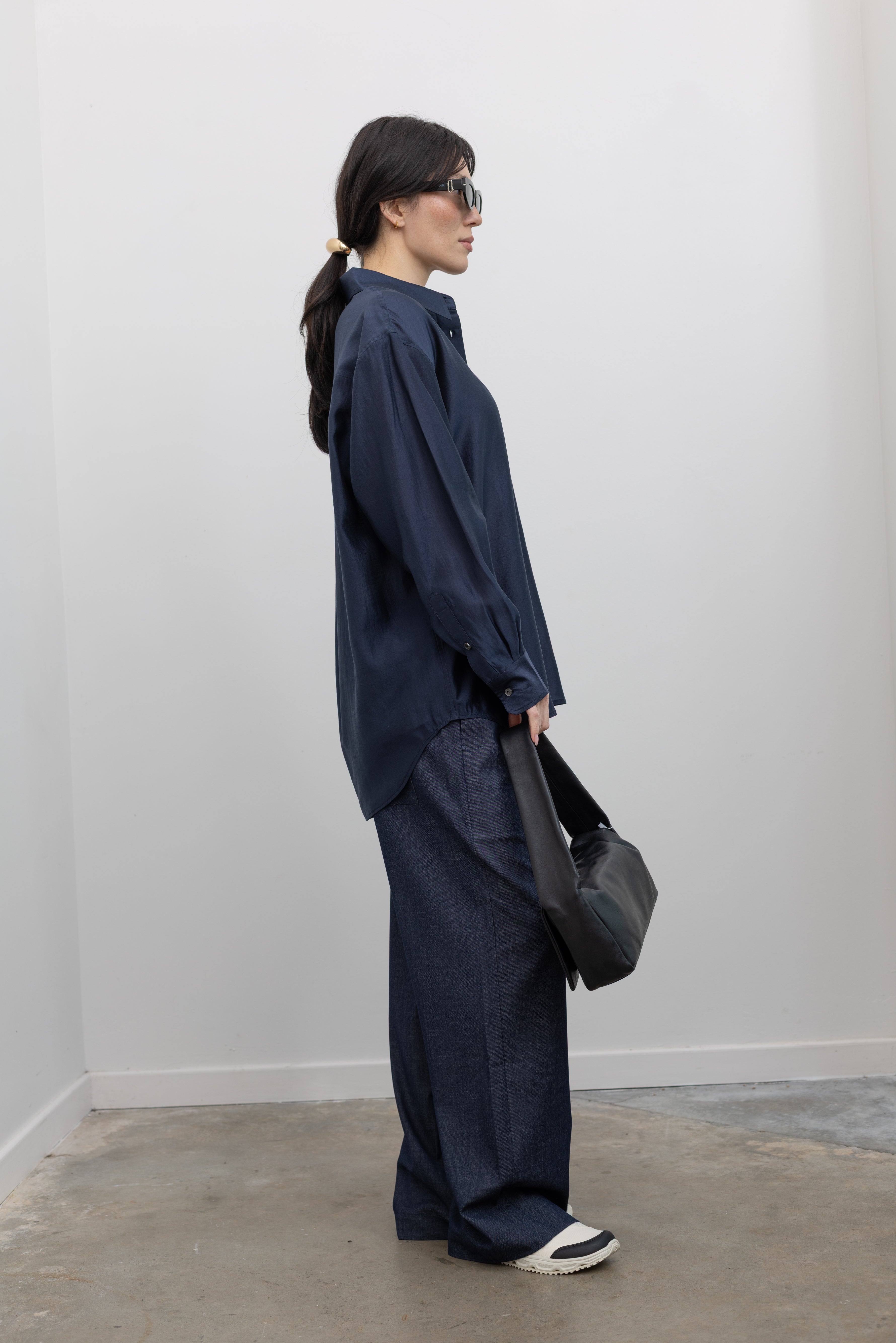 Full length side view of a woman wearing an elevated weekend staple in a Mijeong Park navy blue semi-sheer oversized shirt featuring a flattering off-center button front, long sleeves, and a classic sharp collar paired with Mijeong Park lightweight dark wash denim drawstring pants, Salomon slide sneakers, and a leather St. Agni bag at an Alas Eius store. 