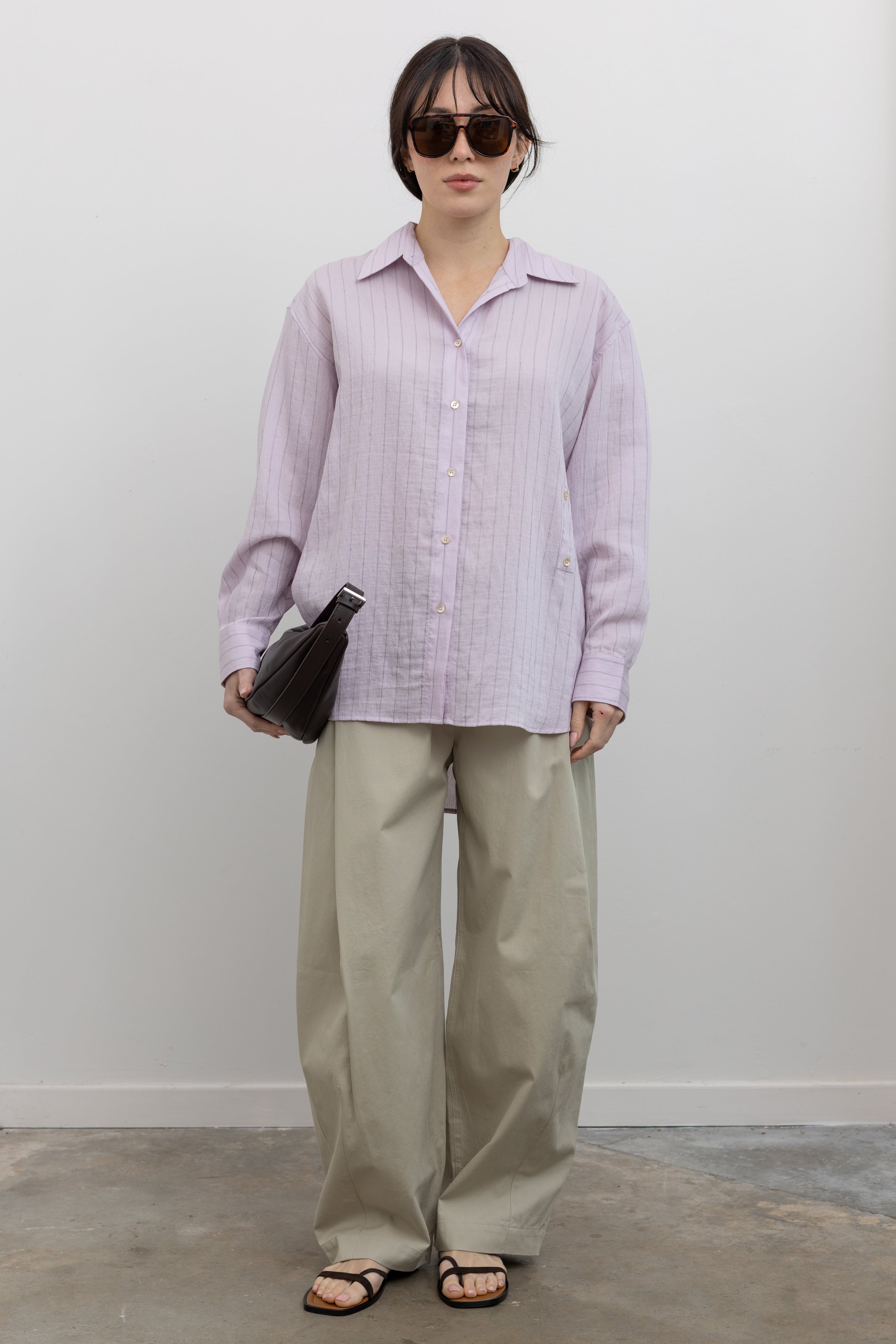 Full length front view of a woman wearing chic spring fashion in a Mijeong Park oversized soft pink pinstripe button down shirt featuring a classic collar, long sleeves, and an off center button down front paired with an elevated classic khaki baggy curved leg pant with strappy sandals at an Alas Eius store. 