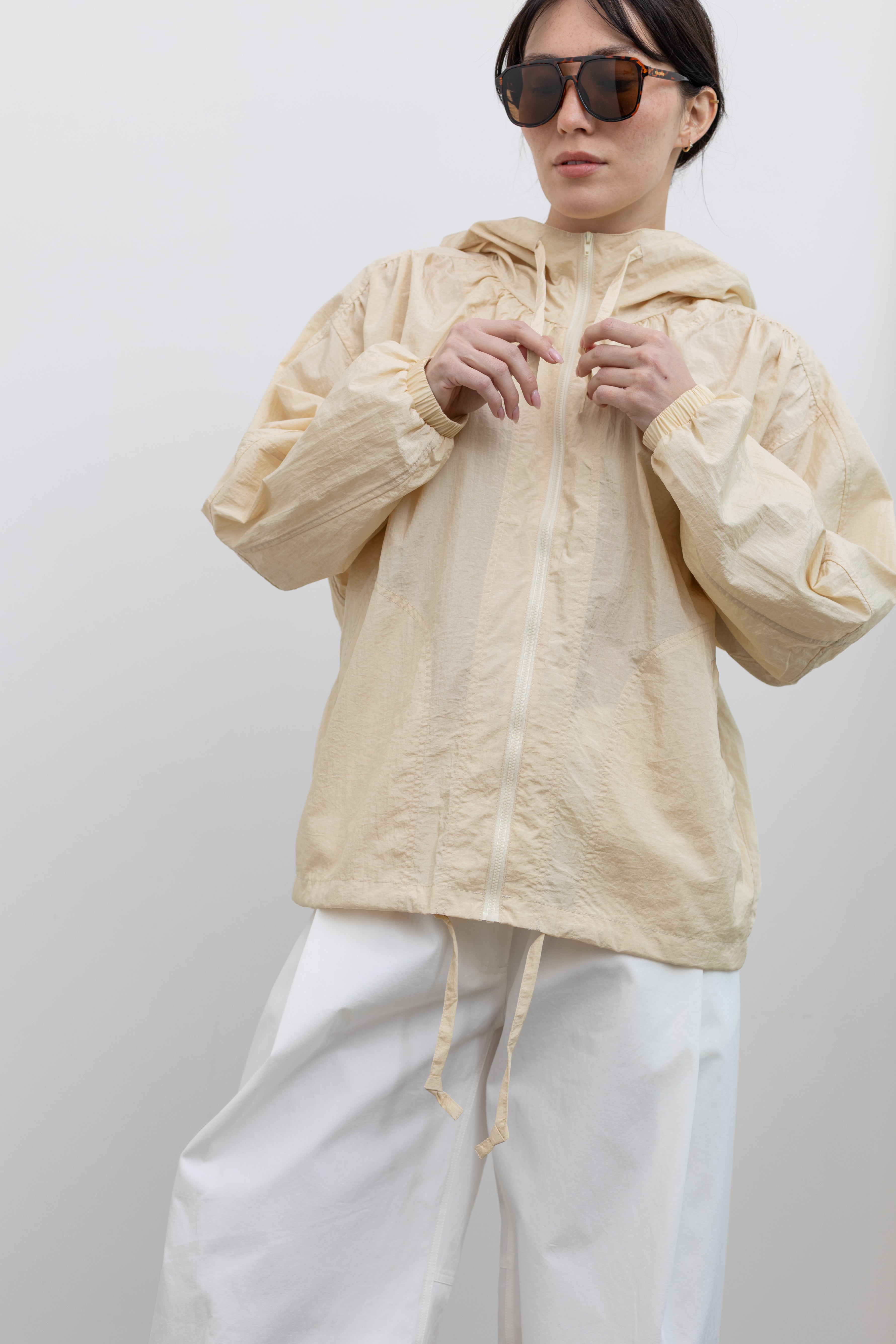 Detailed front view of a woman wearing an elevated 90's inspired outerwear staple in a Mijeong Park cream colored nylon shirred anorak jacket featuring an oversized fit, front pockets, hood, and a zipper closure paired with white curved leg pants at an Alas Eius store. 