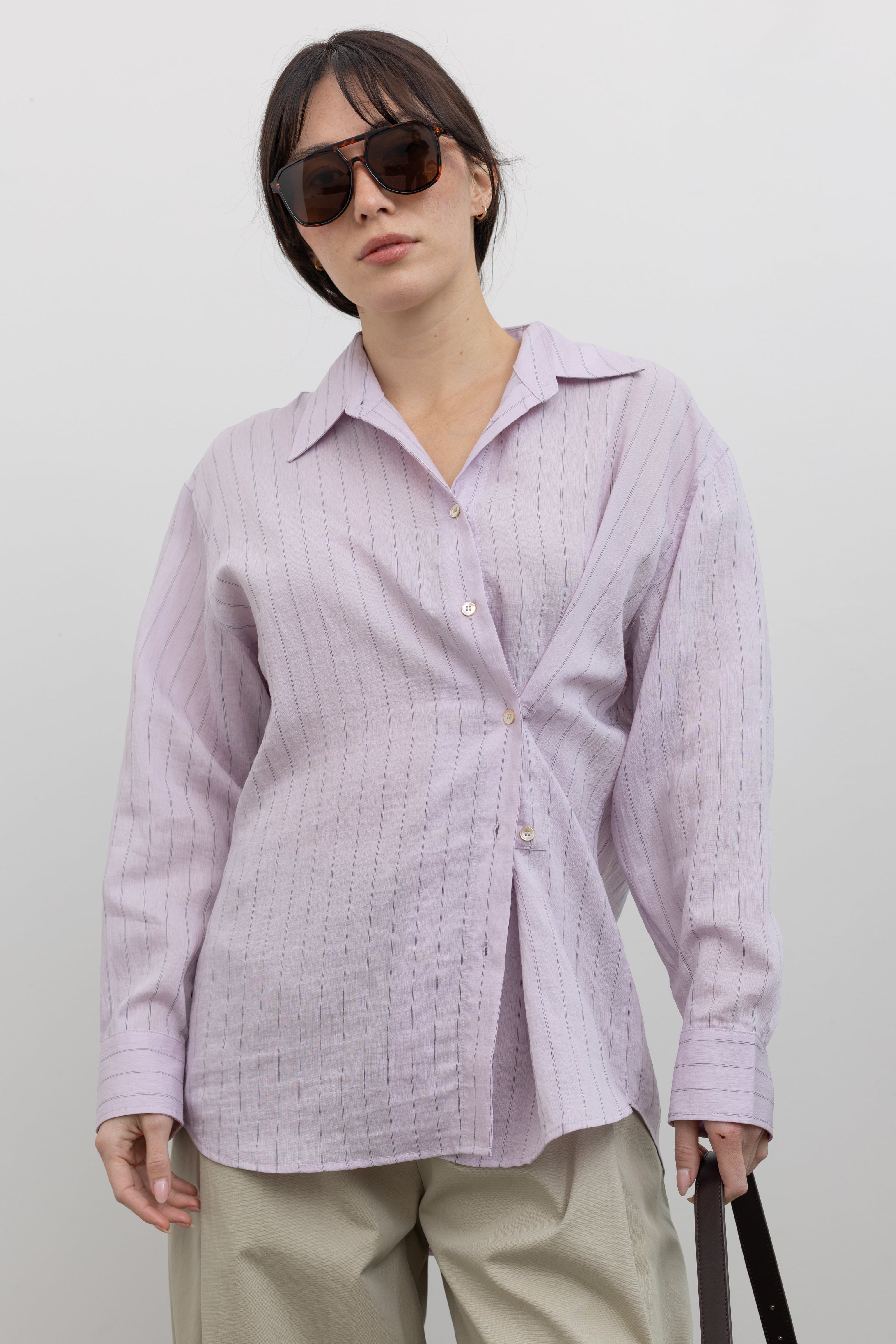 Close-up front view of a woman wearing elevated 90's inspired capsule wardrobe staple in a Mijeong Park oversized soft pink pinstripe button down shirt featuring a classic collar, long sleeves, and an off center button down front at an Alas Eius store. 