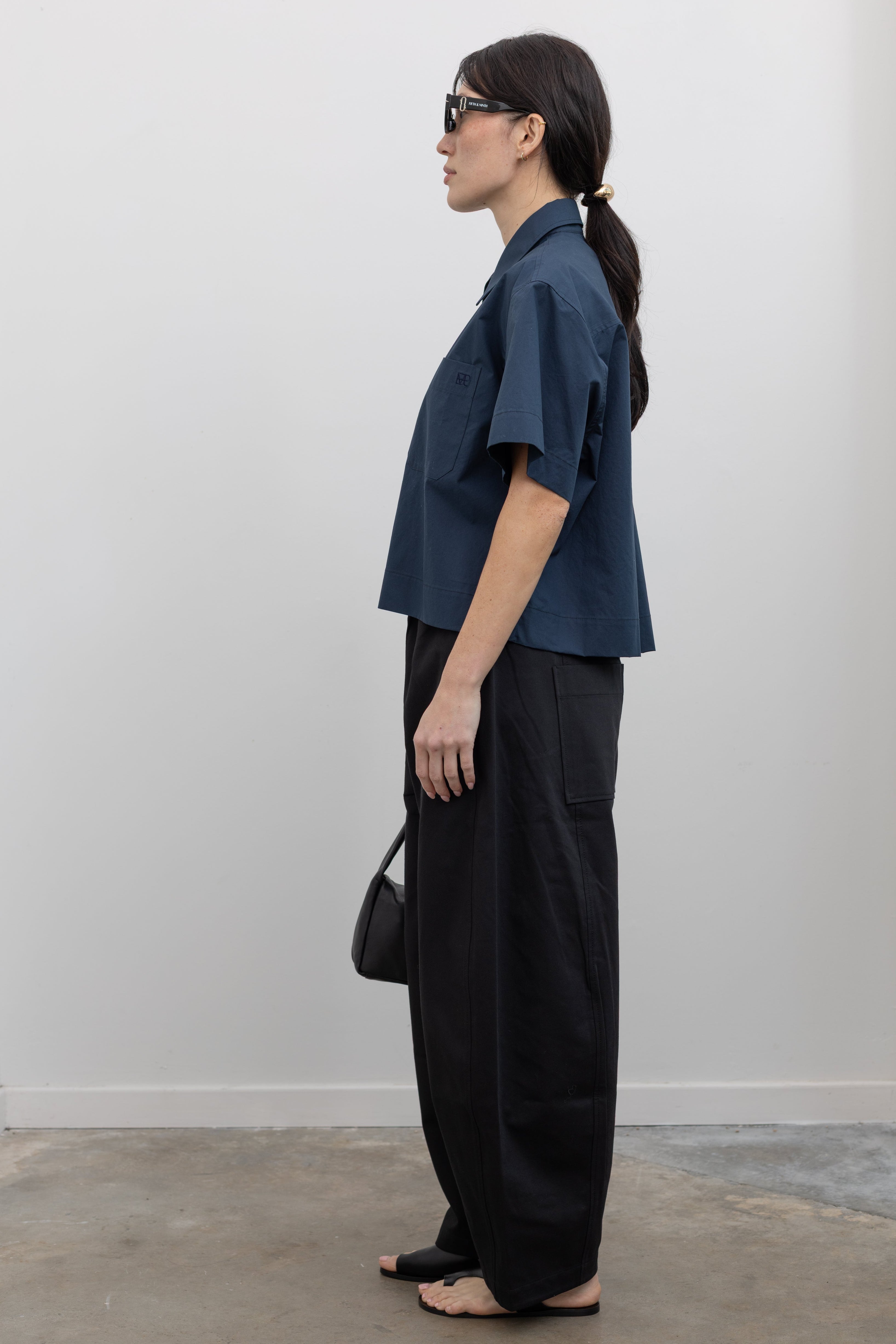 Full length side view of contemporary womenswear spring fashion in Mijeong Park black baggy balloon pants with curved outseams, a relaxed fit, elastic waistband, and back patch pockets paired with a structured navy blue short sleeve cropped hem button down shirt and a black leather St. Agni bag at an Alas Eius store. 