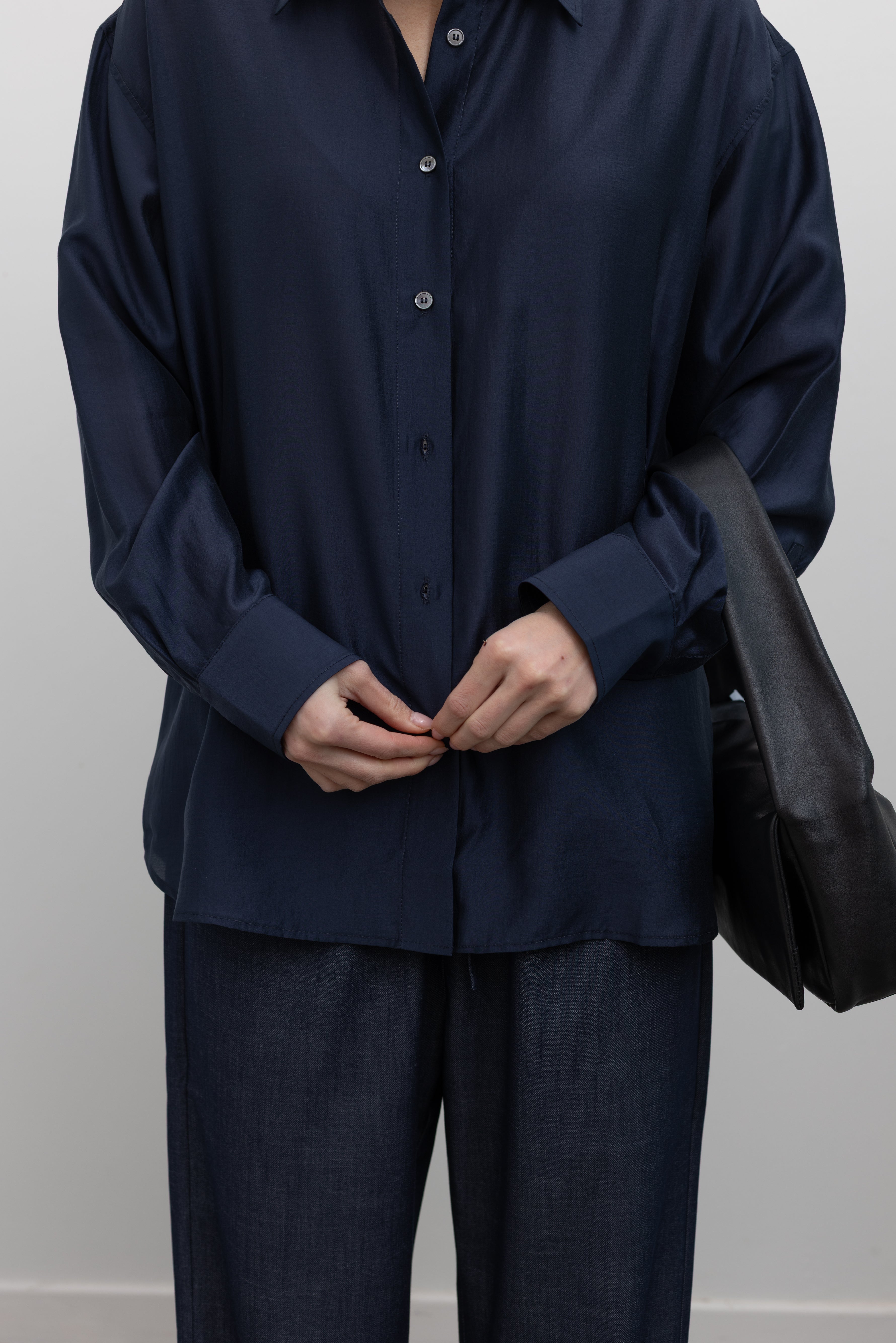 Close-up front view of a woman wearing a modern minimalist staple in a Mijeong Park navy blue semi-sheer oversized shirt featuring a flattering off-center button front, long sleeves, and a classic sharp collar paired with Mijeong Park lightweight dark wash denim drawstring pants, and a leather St. Agni bag at an Alas Eius store. 