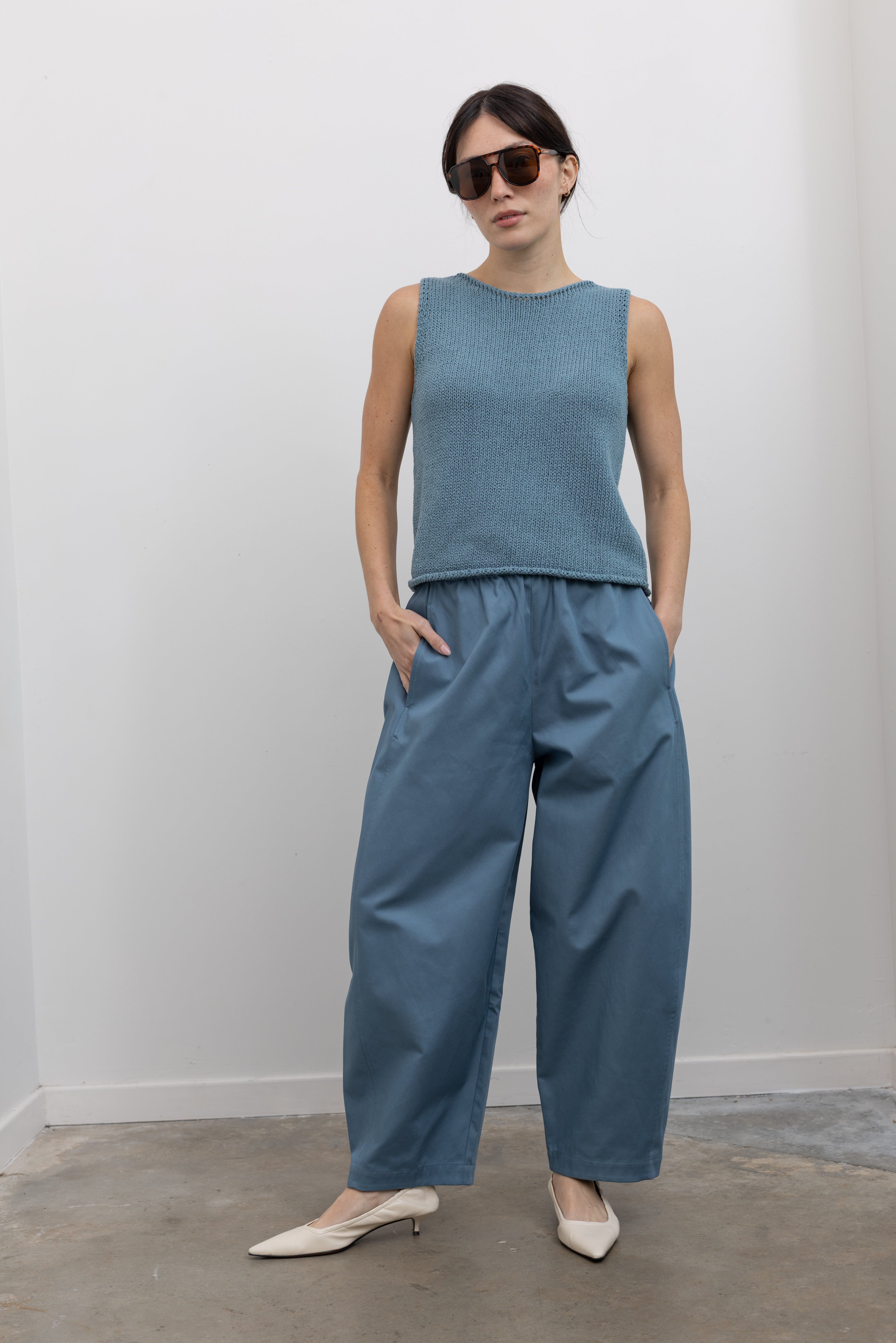 Full length front view of a woman wearing spring fashion in a Mijeong Park light blue boat neck knit sweater tank top featuring rolled edges around the neckline and hem paired with light blue baggy barrel pants at an Alas Eius store. 