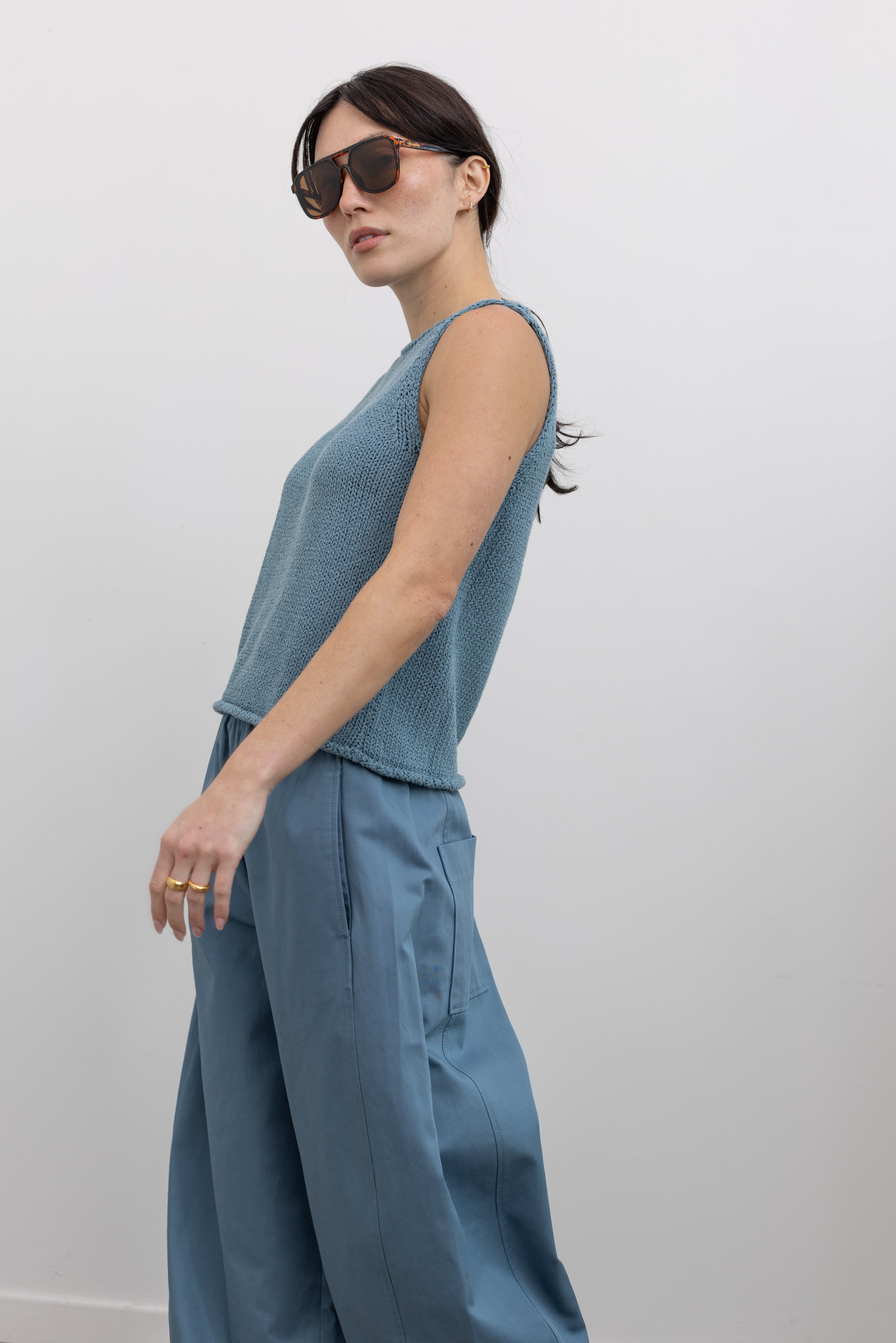 Side view of a woman in motion wearing elevated spring relaxed fashion in a Mijeong Park light blue boat neck knit sweater tank top featuring rolled edges around the neckline and hem at an Alas Eius store. 