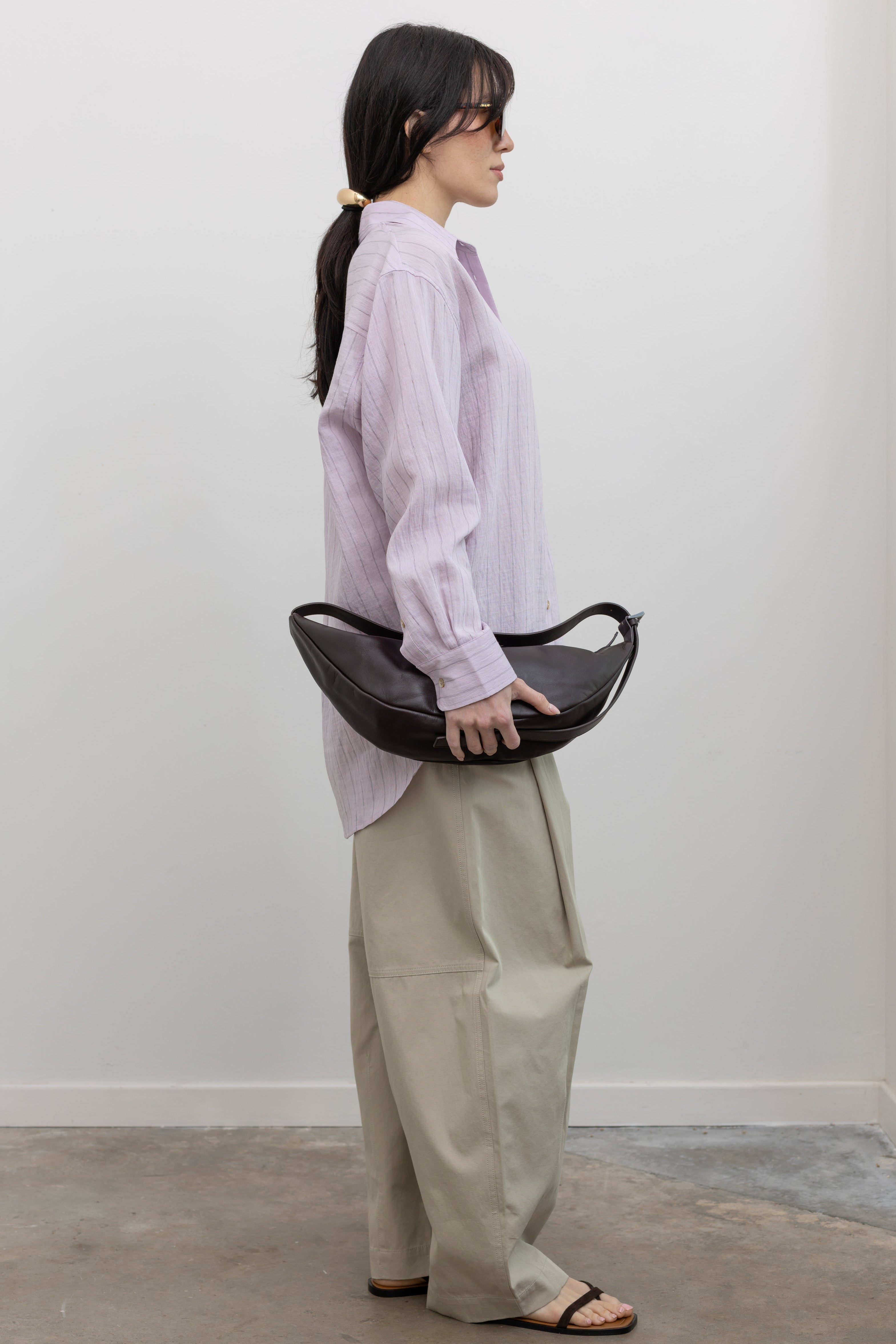 Full length side view of a woman wearing modern minimalist spring fashion in a Mijeong Park oversized soft pink pinstripe button down shirt featuring a classic collar, long sleeves, and an off center button down front paired with an elevated classic khaki baggy curved leg pant with strappy sandals at an Alas Eius store. 