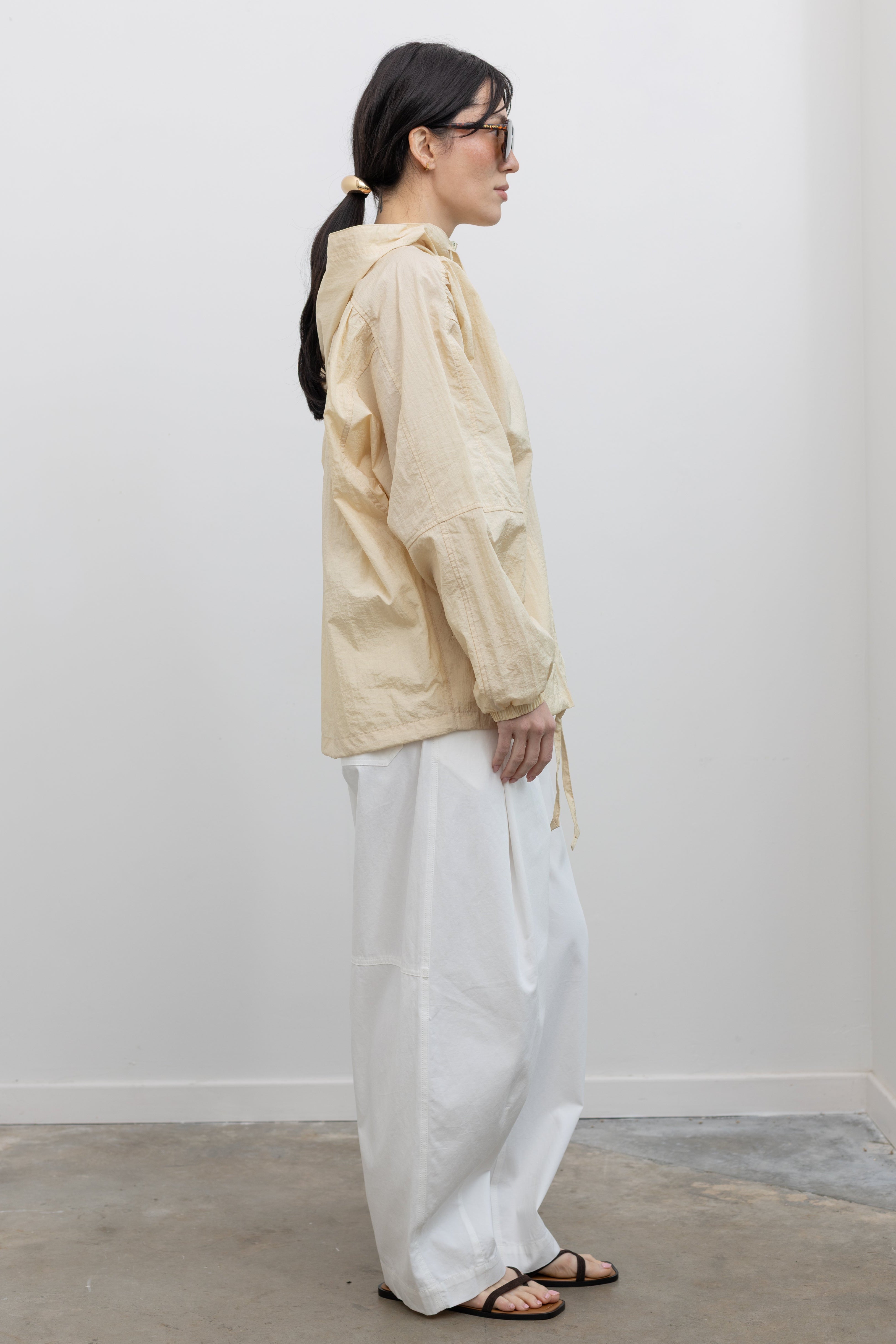 Full length side view of a woman wearing an elevated 90's inspired outerwear staple in a Mijeong Park cream colored nylon shirred anorak jacket featuring an oversized fit, front pockets, hood, and a zipper closure paired with white curved leg pants at an Alas Eius store. 