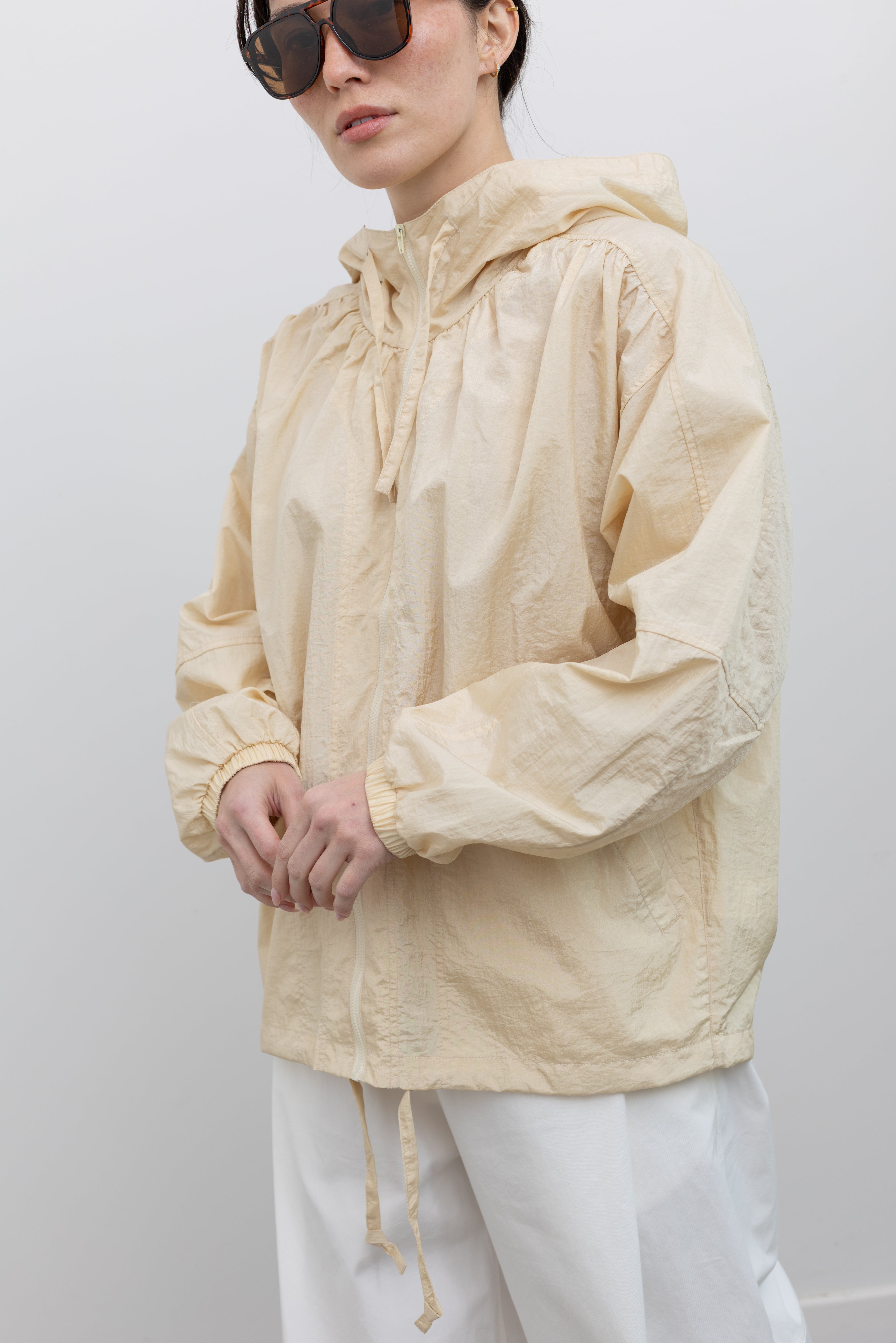 Close-up 3/4 view of a woman wearing an travel outerwear essential in a Mijeong Park cream colored nylon shirred anorak jacket featuring an oversized fit, front pockets, hood, and a zipper closure paired with white curved leg pants at an Alas Eius store. 