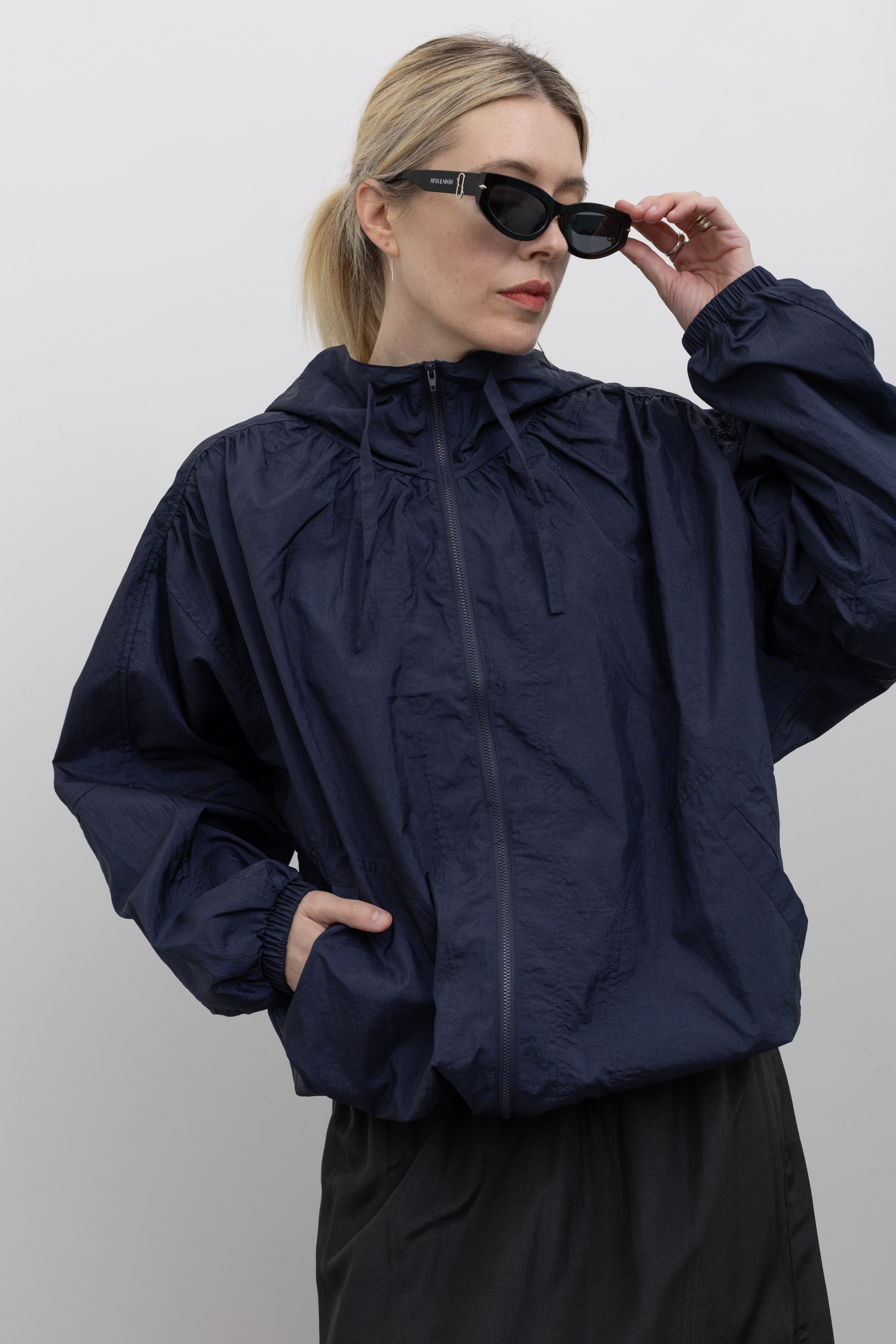 Close-up front view of a woman posing with sunglasses wearing an versatile outerwear staple in a  Mijeong Park navy blue nylon shirred anorak jacket featuring an oversized fit, front pockets, hood, and a zipper closure at an Alas Eius store. 