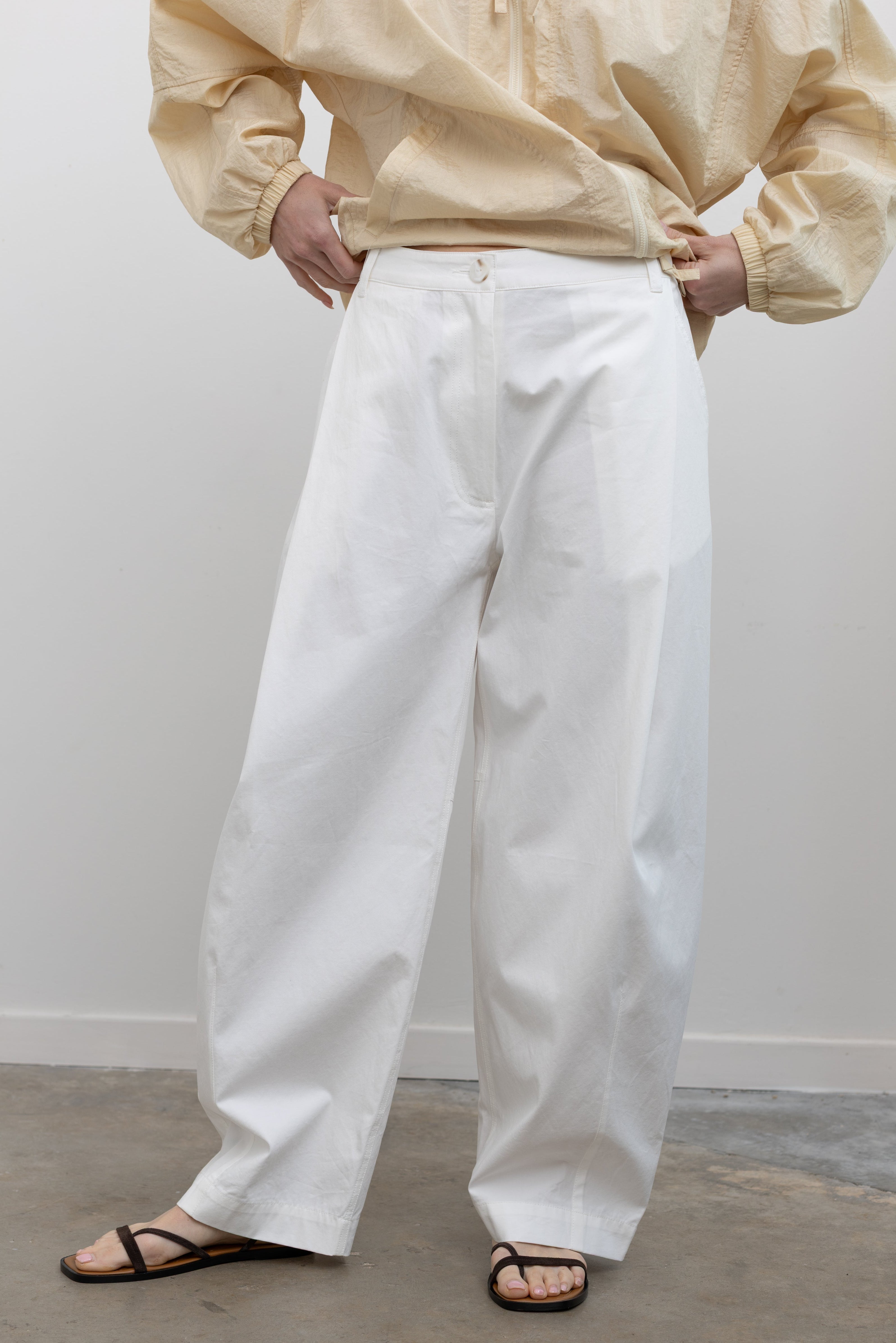 Close-up front view of a woman standing wearing spring fashion in Mijeong Park white 100% cotton barrel leg pants featuring curved outseams that create architectural volume and a partially elasticated waistband and belt loops for a tailored look at an Alas Eius store. 