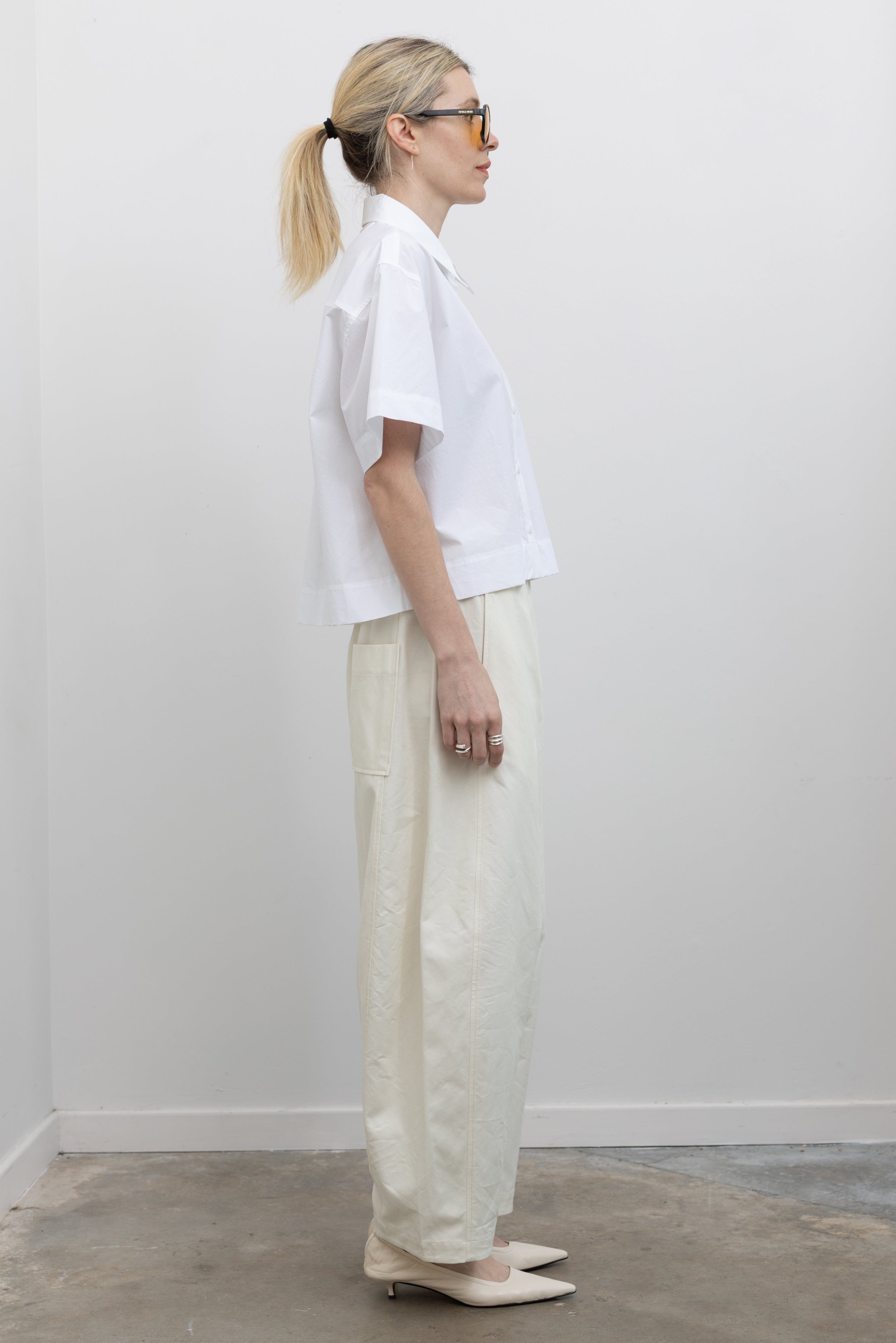 Full length side view of a woman wearing spring minimalist fashion in Mijeong Park cream colored cropped balloon pants with curved outseams, 
a relaxed fit, mid-rise waist, and a comfortable elasticated band paired with a Mijeong Park crisp white cotton oversized cropped collared shirt with a front pocket at an Alas Eius store. 