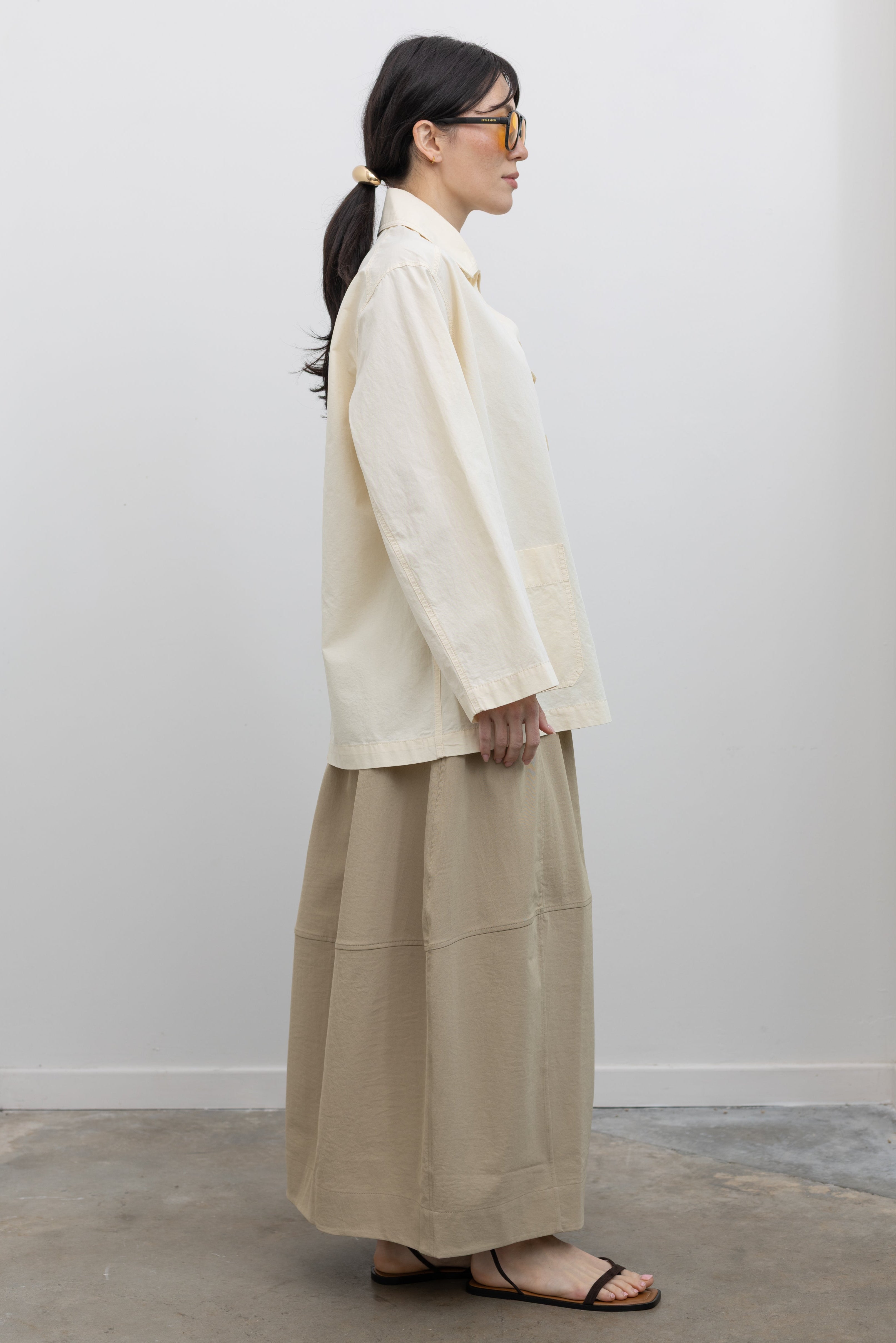 Full length side view of a woman standing wearing chic and modest Spring fashion in a Mijeong Park beige balloon midi skirt paired with a cream colored linen blend work jacket featuring a relaxed fit, two front patch pockets and a button down front closure for versatile styling at an Alas Eius store. 