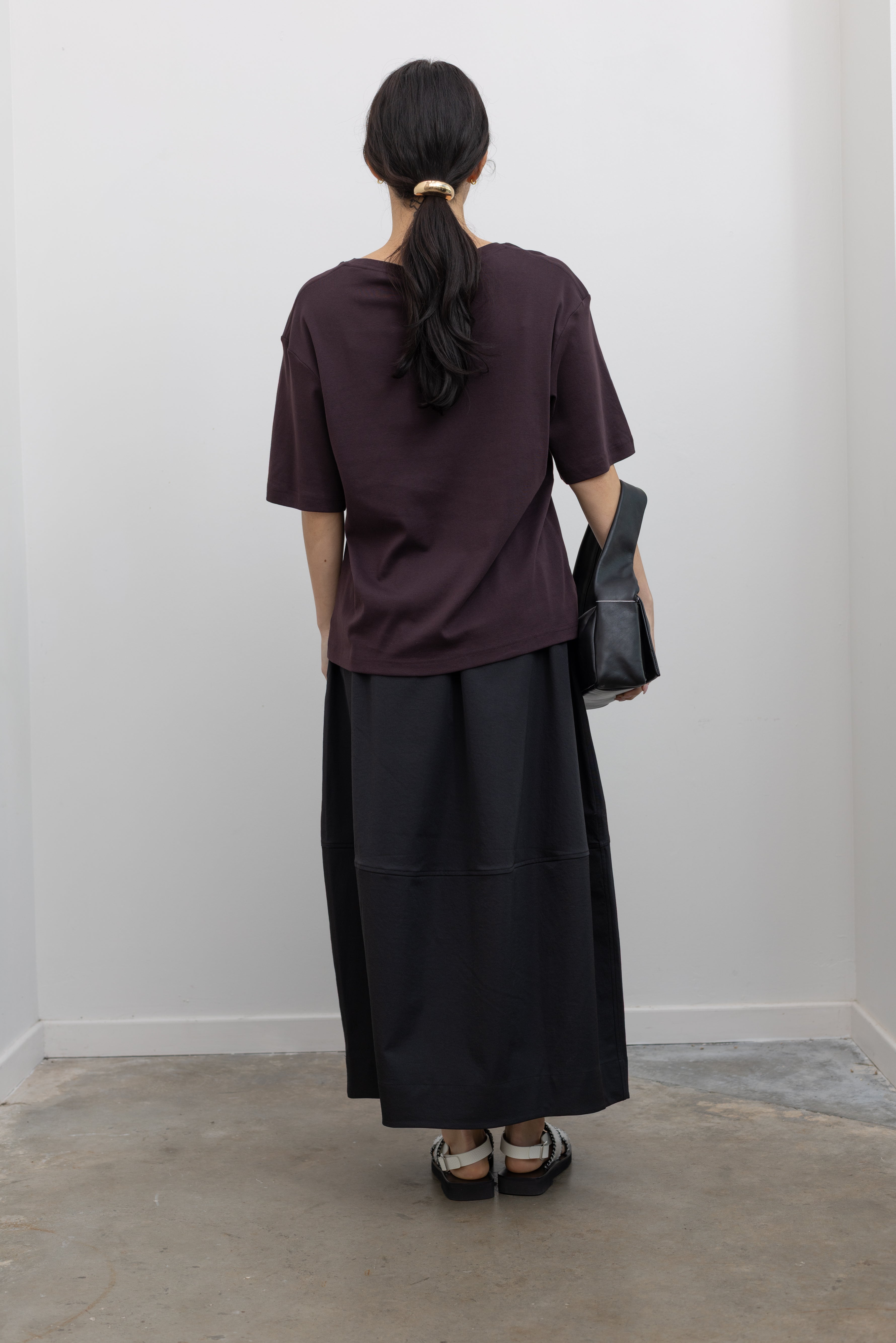 Full length back view of a woman standing wearing contemporary womenswear spring fashion in a Mijeong Park dark brown oversized t-shirt with a black voluminous balloon midi-to-maxi silhouette skirt featuring an elastic waistband paired with silver metallic slip-on sandals and a sleek St. Agni black leather shoulder bag at an Alas Eius store. 