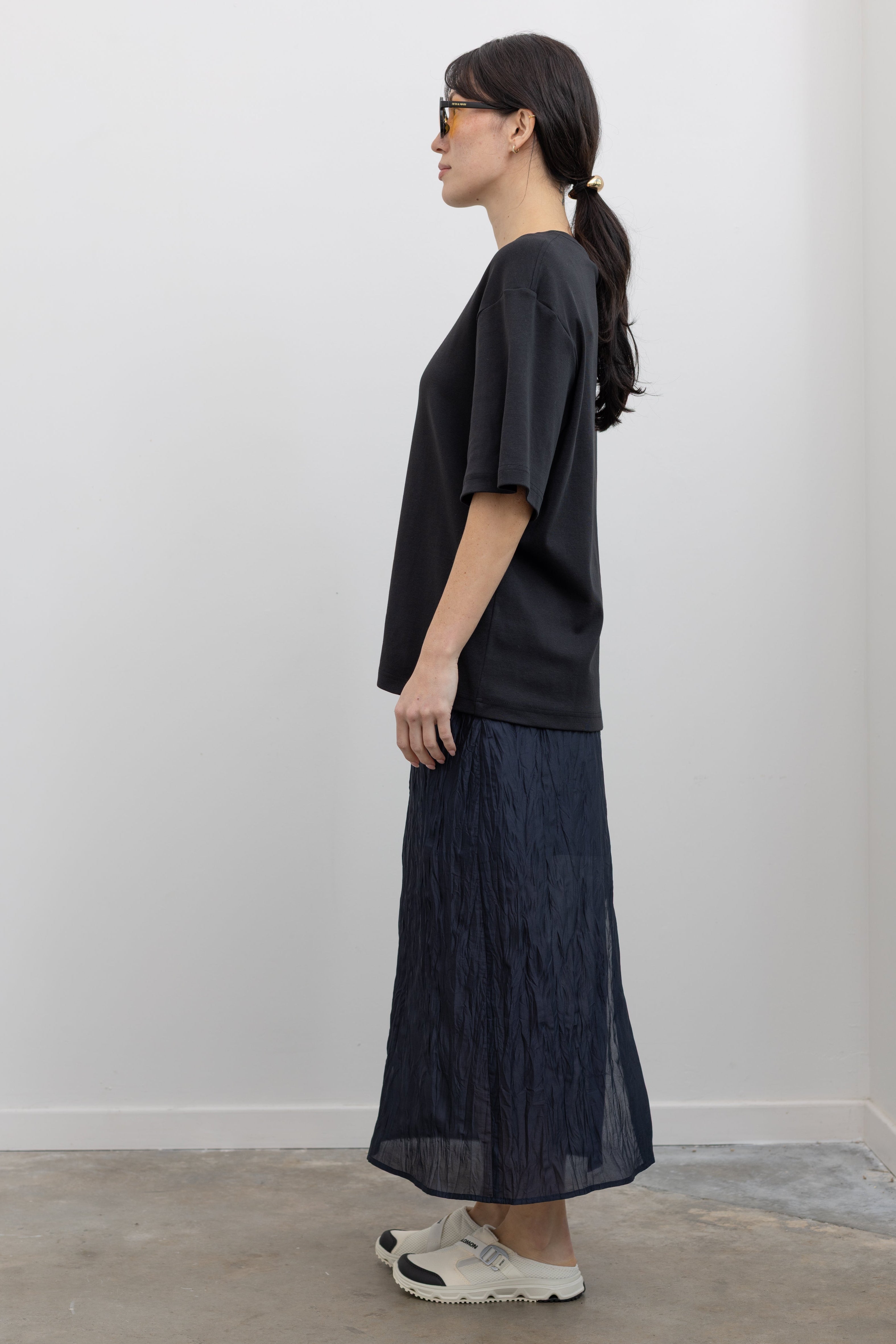 Full length side view of a woman standing wearing spring streetwear fashion in a Mijeong Park navy blue lightweight, semi-sheer crinkled fabric lined midi skirt paired with a Mijeong Park black oversized t-shirt and Salomon sneakers at an Alas Eius store. 