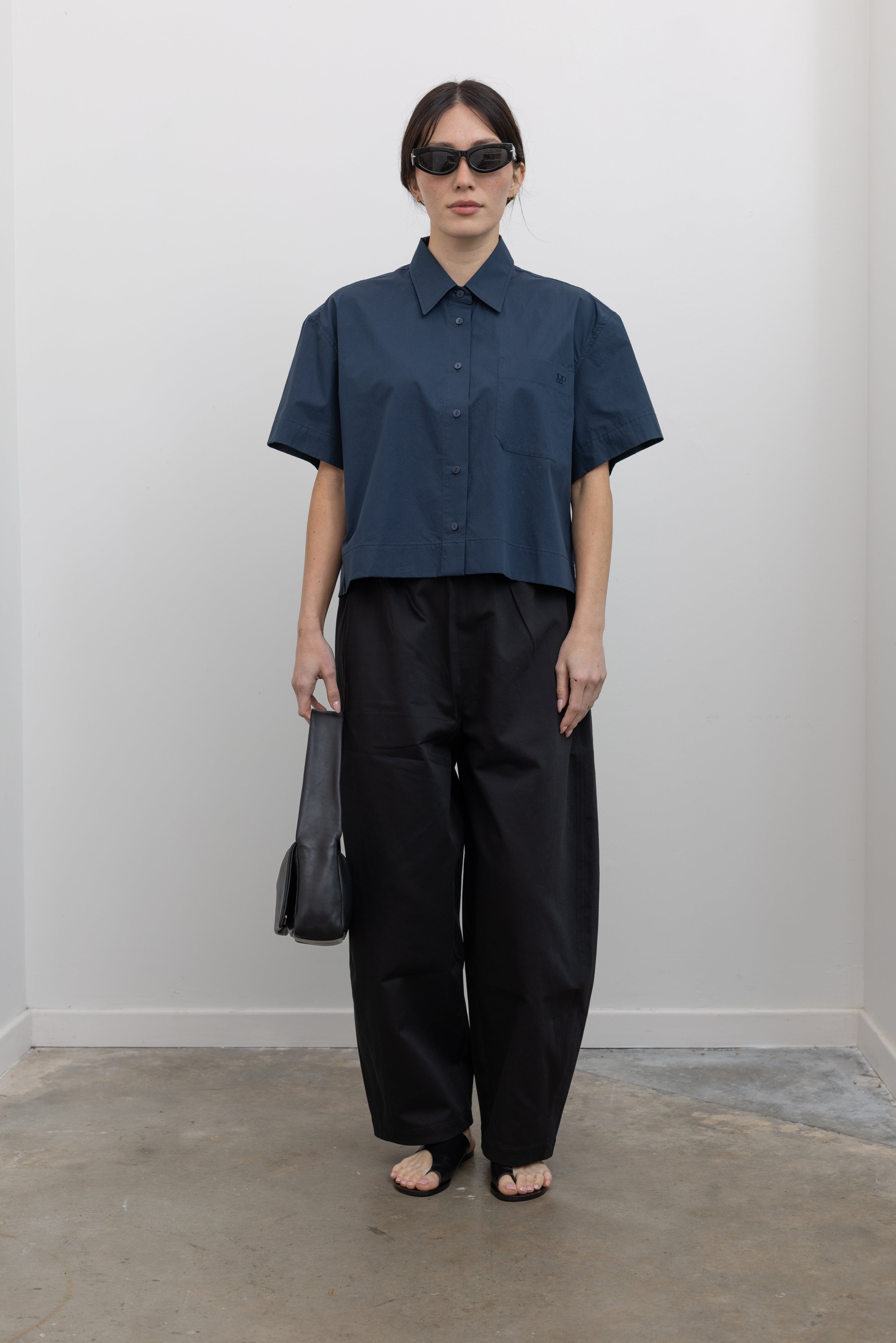 Full length front view of a woman posing with sunglasses wearing modern architectural spring fashion in a Mijeong Park 100% cotton navy blue oversized collared button down shirt featuring a chest patch pocket, a cropped hem, and short sleeves paired with an elevated classic wardrobe staple in Mijeong Park black baggy curved leg pants at an Alas Eius store.