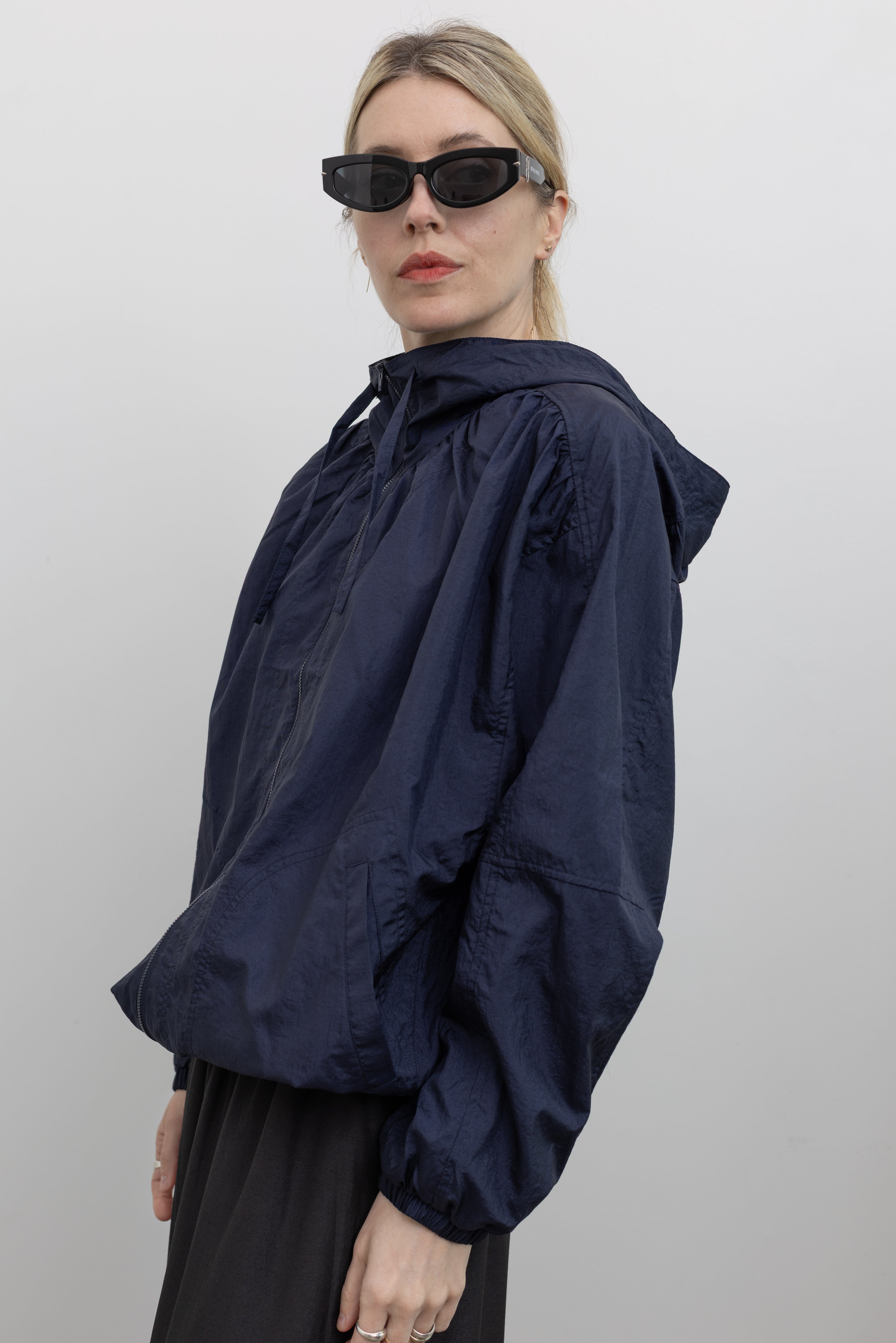 Close-up 3/4 view of a woman wearing an elevated Mijeong Park navy blue nylon shirred anorak jacket featuring an oversized fit, front pockets, hood, and a zipper closure at an Alas Eius store. 