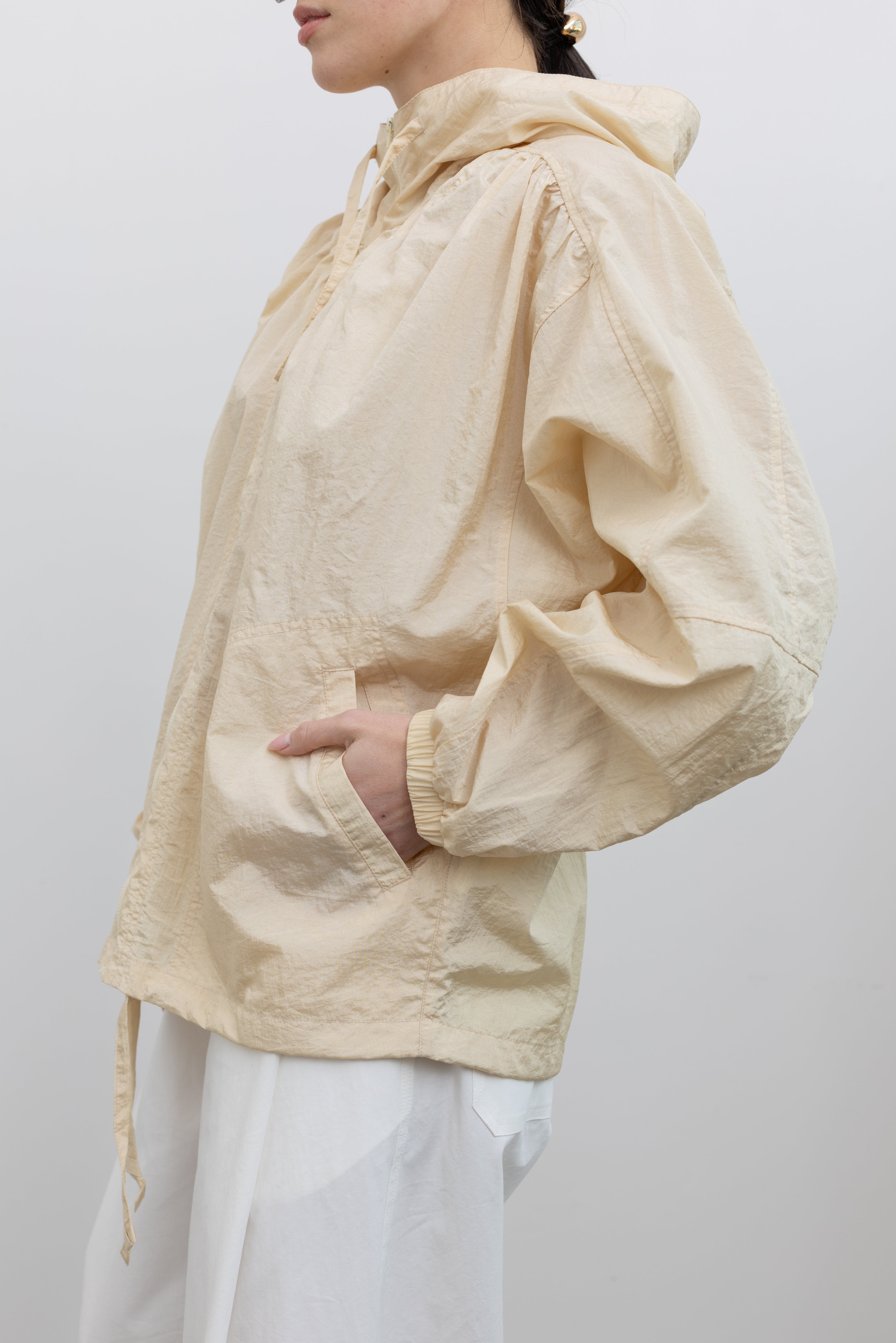 Close-up side view of a woman wearing an elevated outerwear staple in a Mijeong Park cream colored nylon shirred anorak jacket featuring an oversized fit, front pockets, hood, and a zipper closure paired with white curved leg pants at an Alas Eius store. 