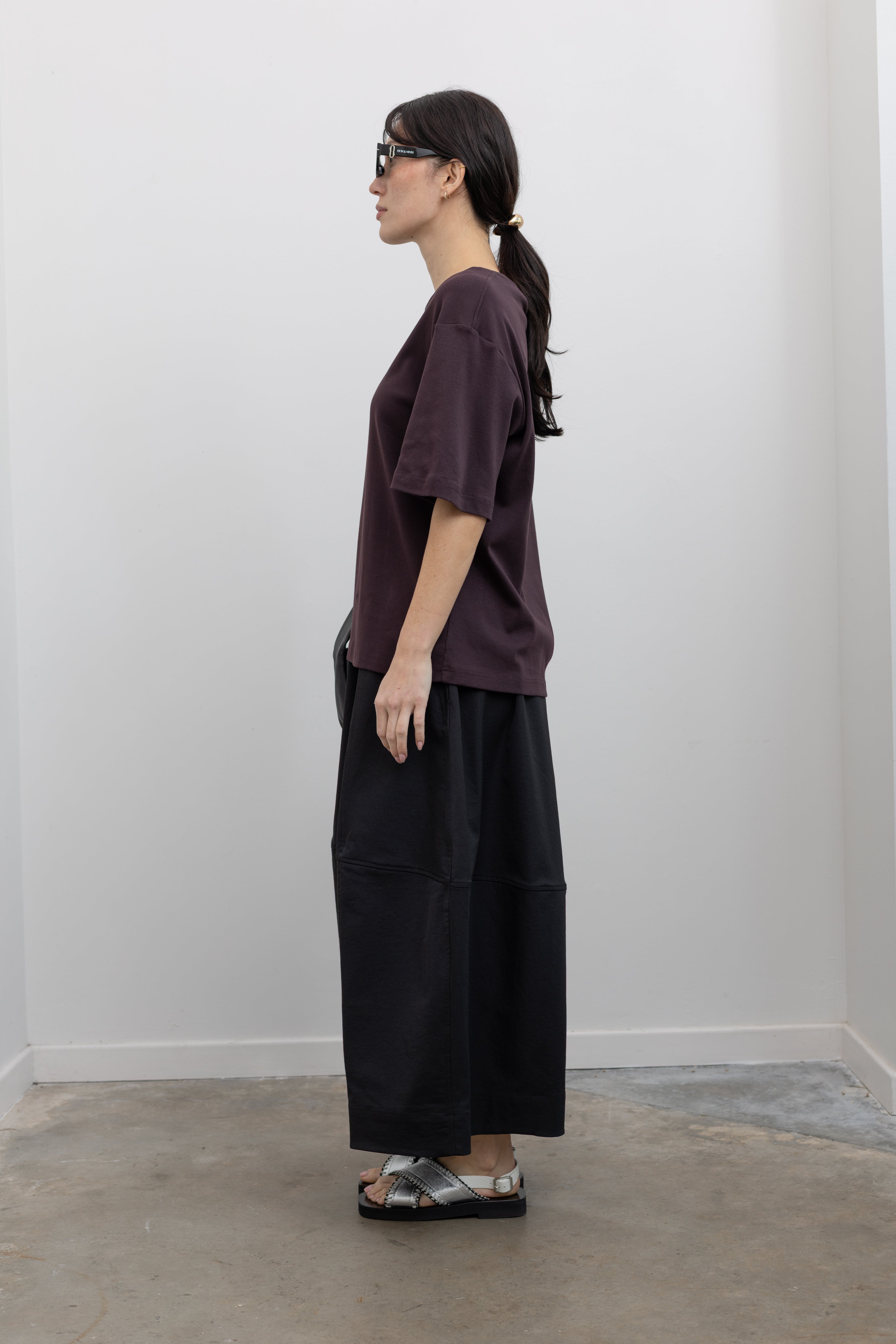 Full length side view of a woman standing wearing contemporary womenswear spring fashion in a Mijeong Park dark brown oversized t-shirt with a black voluminous balloon midi-to-maxi silhouette skirt featuring an elastic waistband paired with silver metallic slip-on sandals and a sleek St. Agni black leather shoulder bag at an Alas Eius store. 