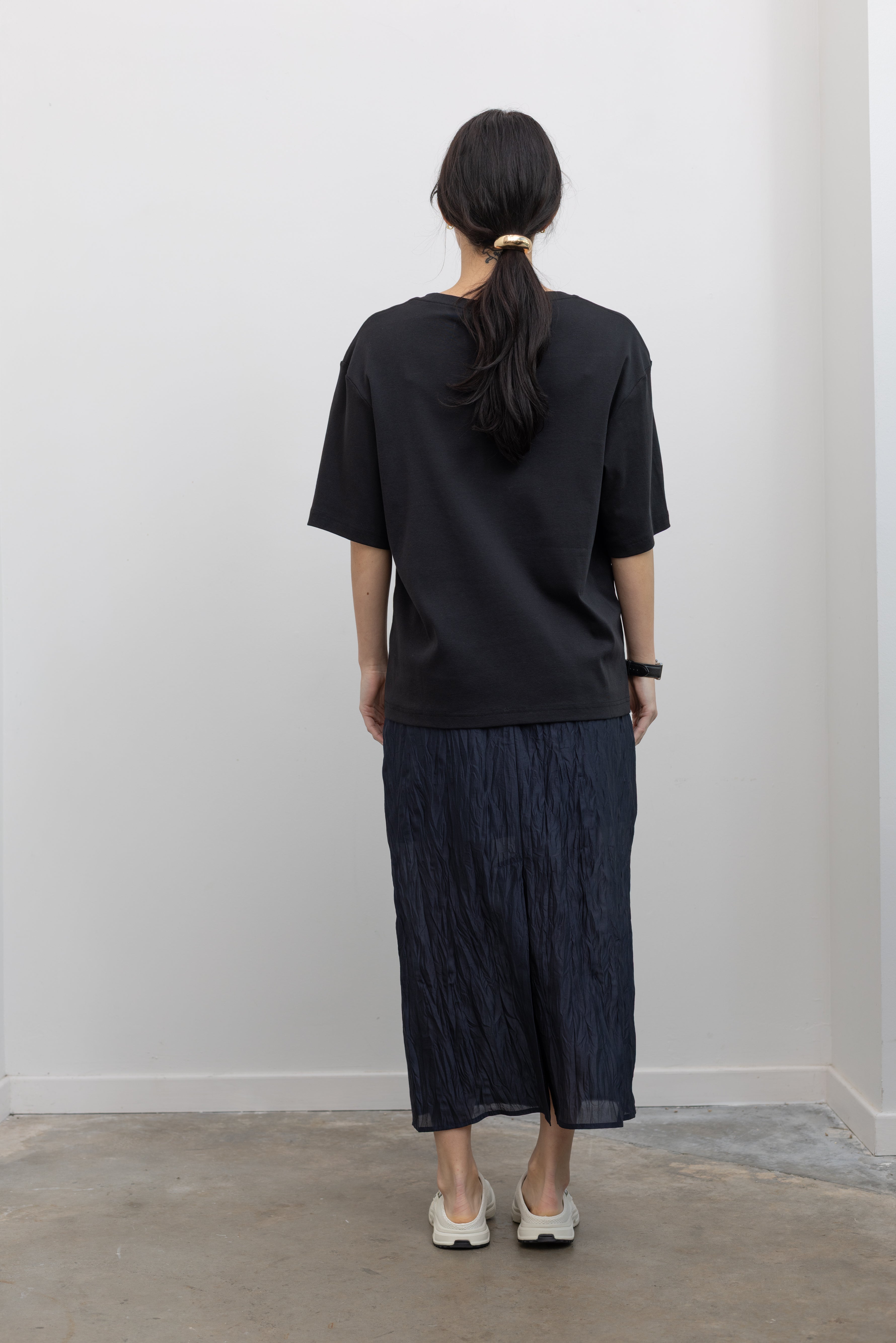 Full length back view of a woman standing wearing elevated essentials spring fashion in a Mijeong Park navy blue lightweight, semi-sheer crinkled fabric lined midi skirt paired with a Mijeong Park black oversized t-shirt and Salomon sneakers at an Alas Eius store. 