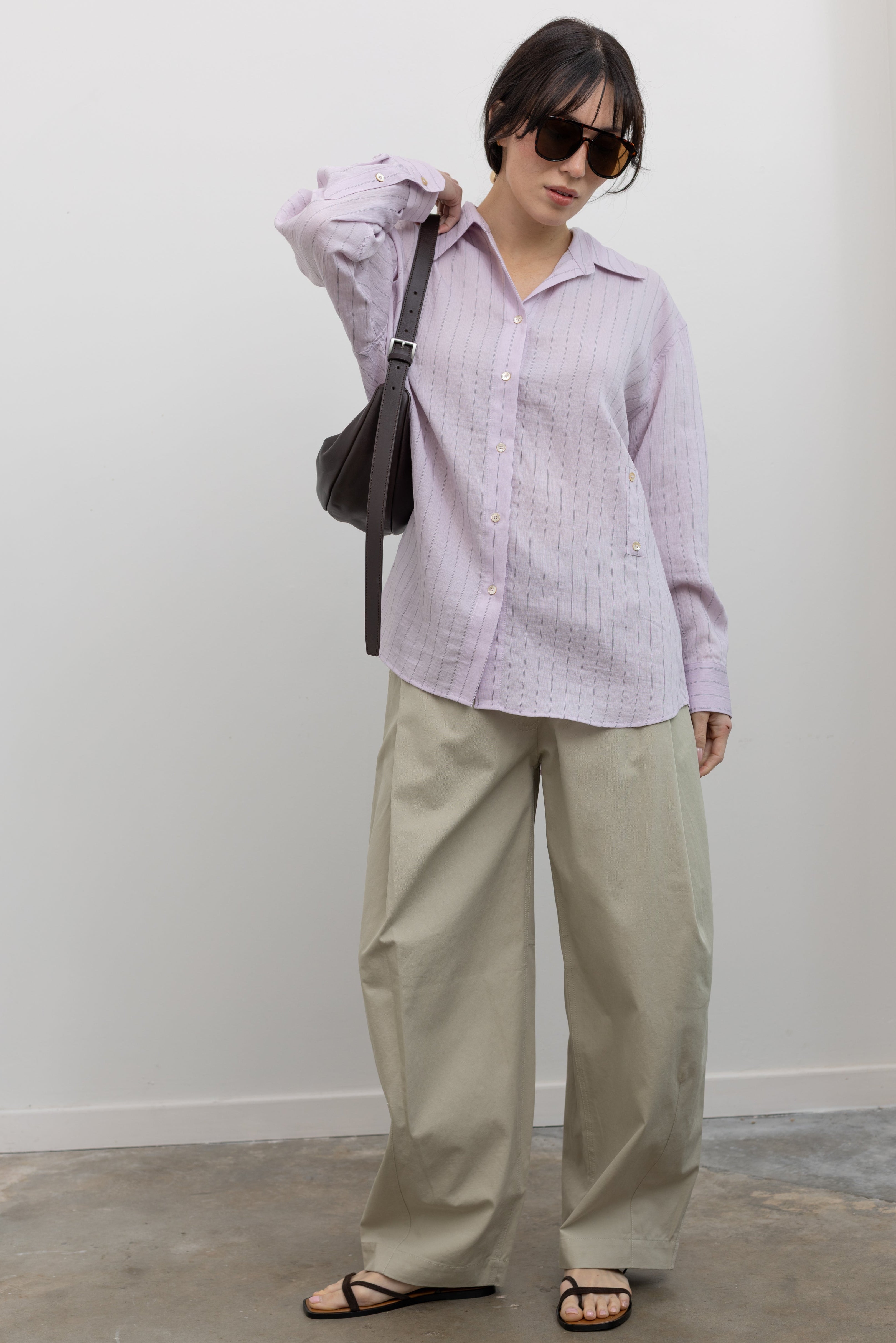 Full length front view of a woman wearing elevated weekend and travel capsule wardrobe staples in a Mijeong Park oversized soft pink pinstripe button down shirt featuring a classic collar, long sleeves, and an off center button down front paired with an elevated classic khaki baggy curved leg pant with strappy sandals at an Alas Eius store. 