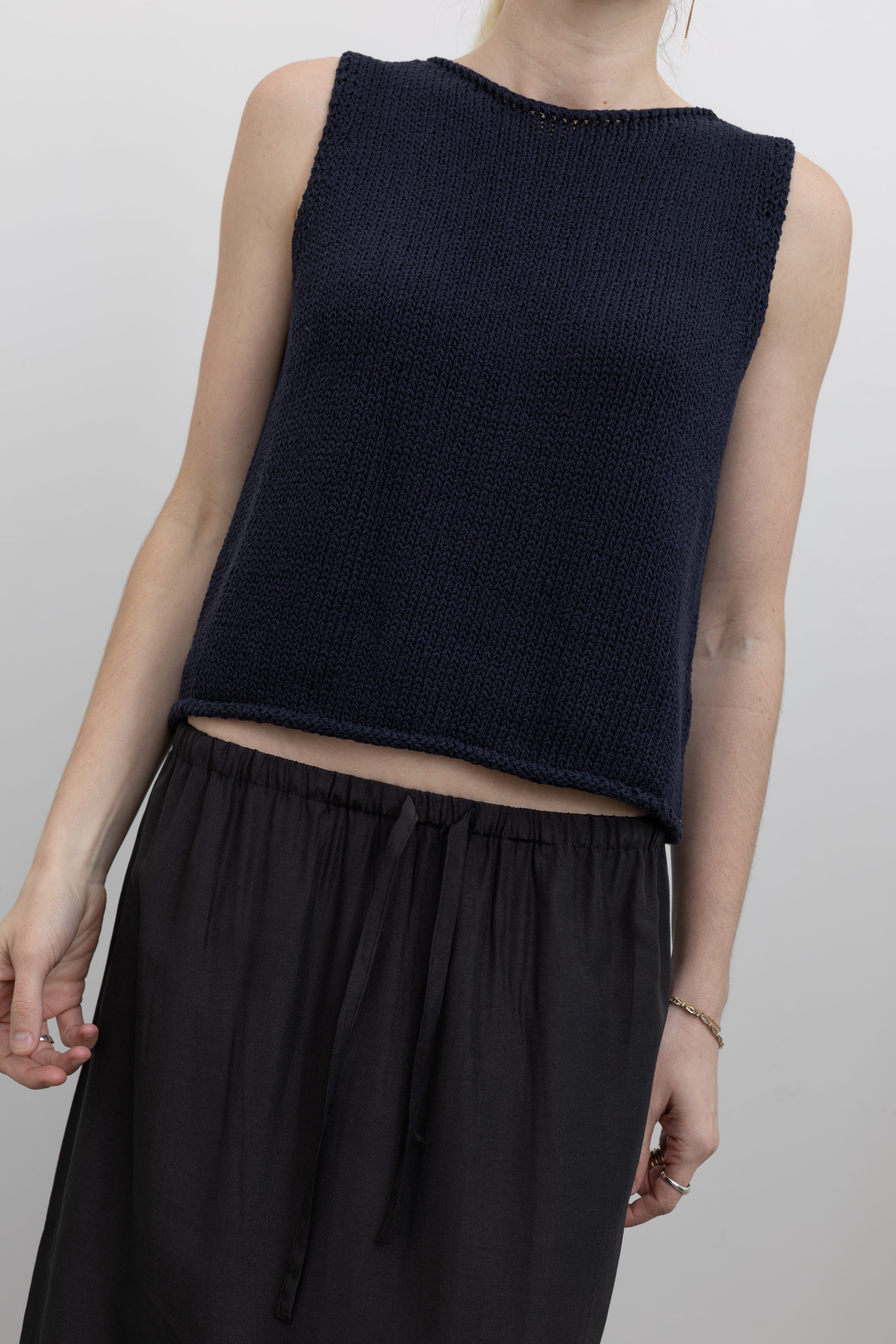 Front view of a woman standing wearing a versatile spring fashion staple in a Mijeong Park navy blue boat neck knit sweater tank top featuring rolled edges around the neckline and hem paired with a Mijeong Park black drawstring waist maxi skirt at an Alas Eius store. 