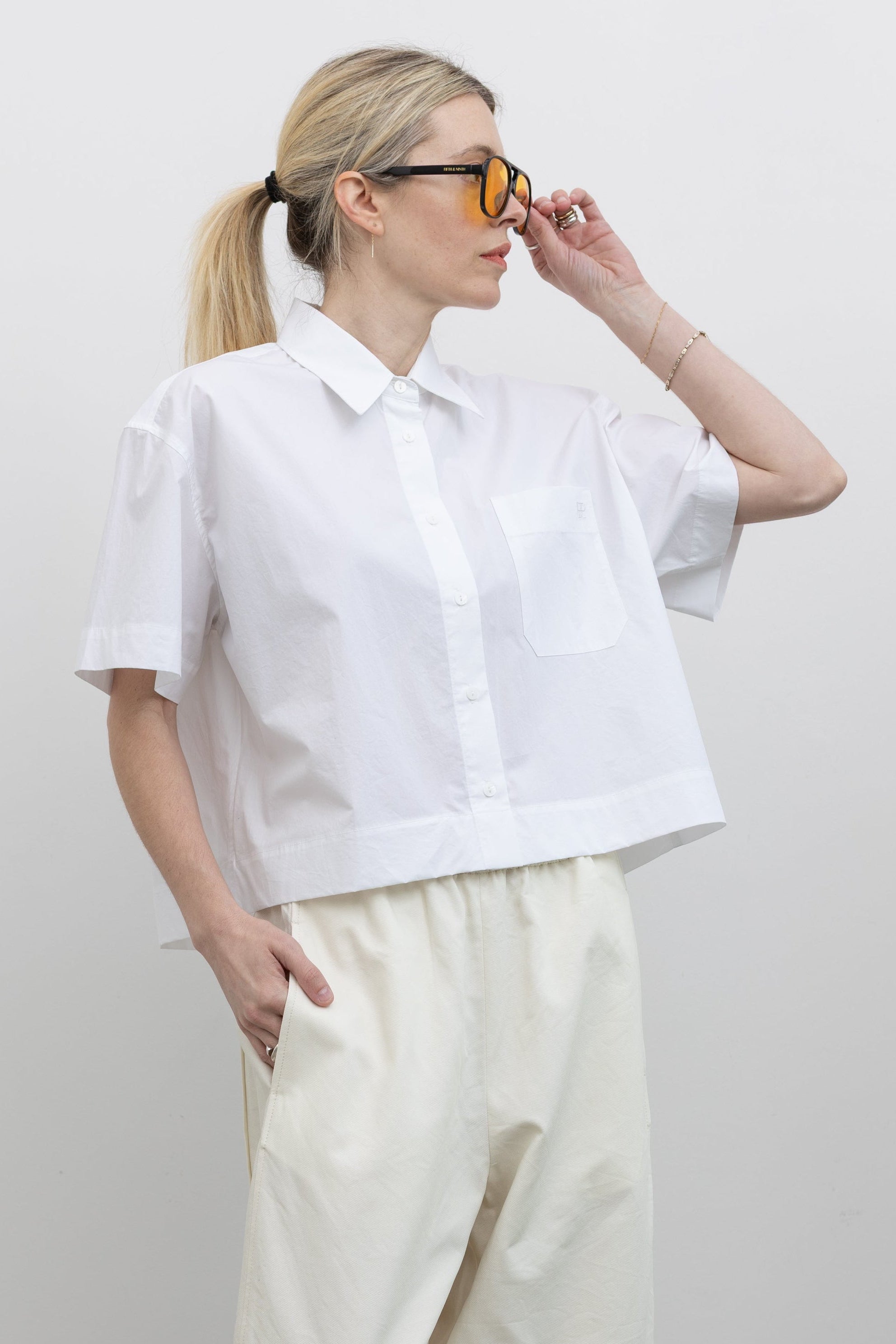 Front view of a woman posing with sunglasses wearing modern minimalist spring fashion in a Mijeong Park 100% cotton white oversized collared shirt featuring a chest patch pocket, a cropped hem, and short sleeves paired with an elevated classic wardrobe staple in Mijeong Park cream baggy curved leg trousers at an Alas Eius store.