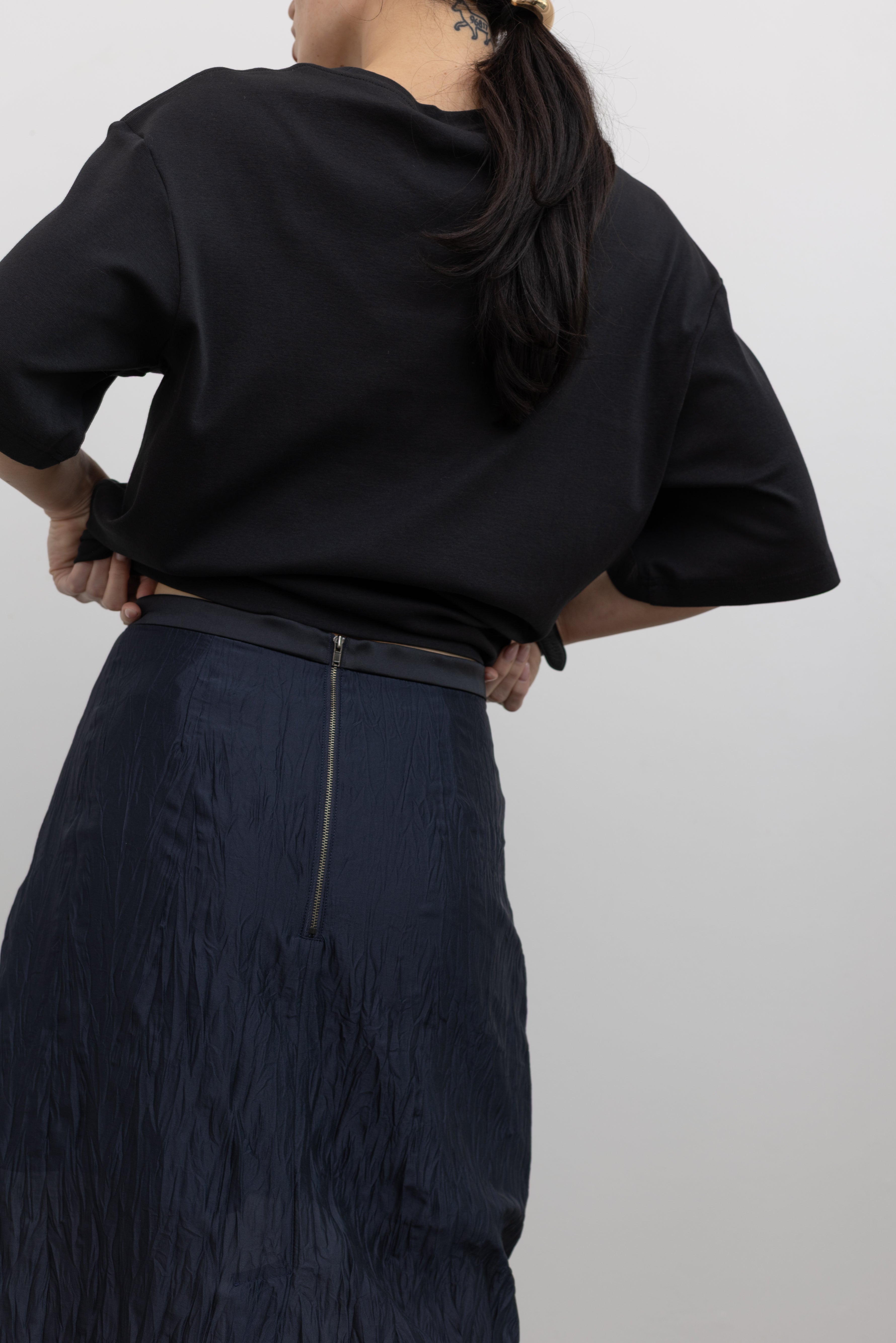 Close-up back view of a woman standing wearing contemporary spring fashion in a Mijeong Park navy blue lightweight, semi-sheer crinkled fabric lined midi skirt paired with a Mijeong Park black oversized t-shirt at an Alas Eius store. 