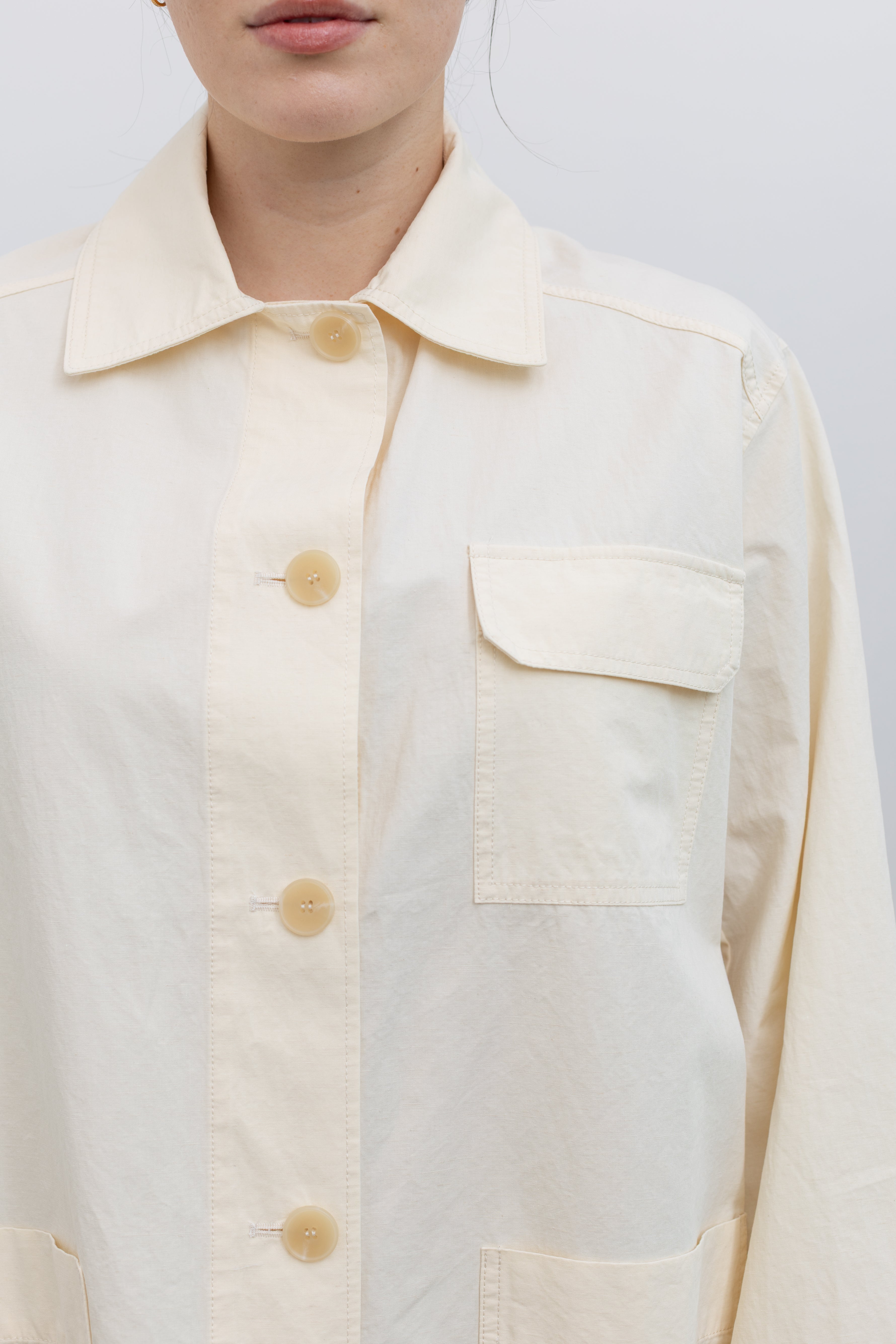 Close-up view of a woman wearing modern and feminine Spring fashion in a Mijeong Park cream colored linen blend work jacket featuring a relaxed fit, two front patch pockets and a button down front closure for versatile styling at an Alas Eius store. 