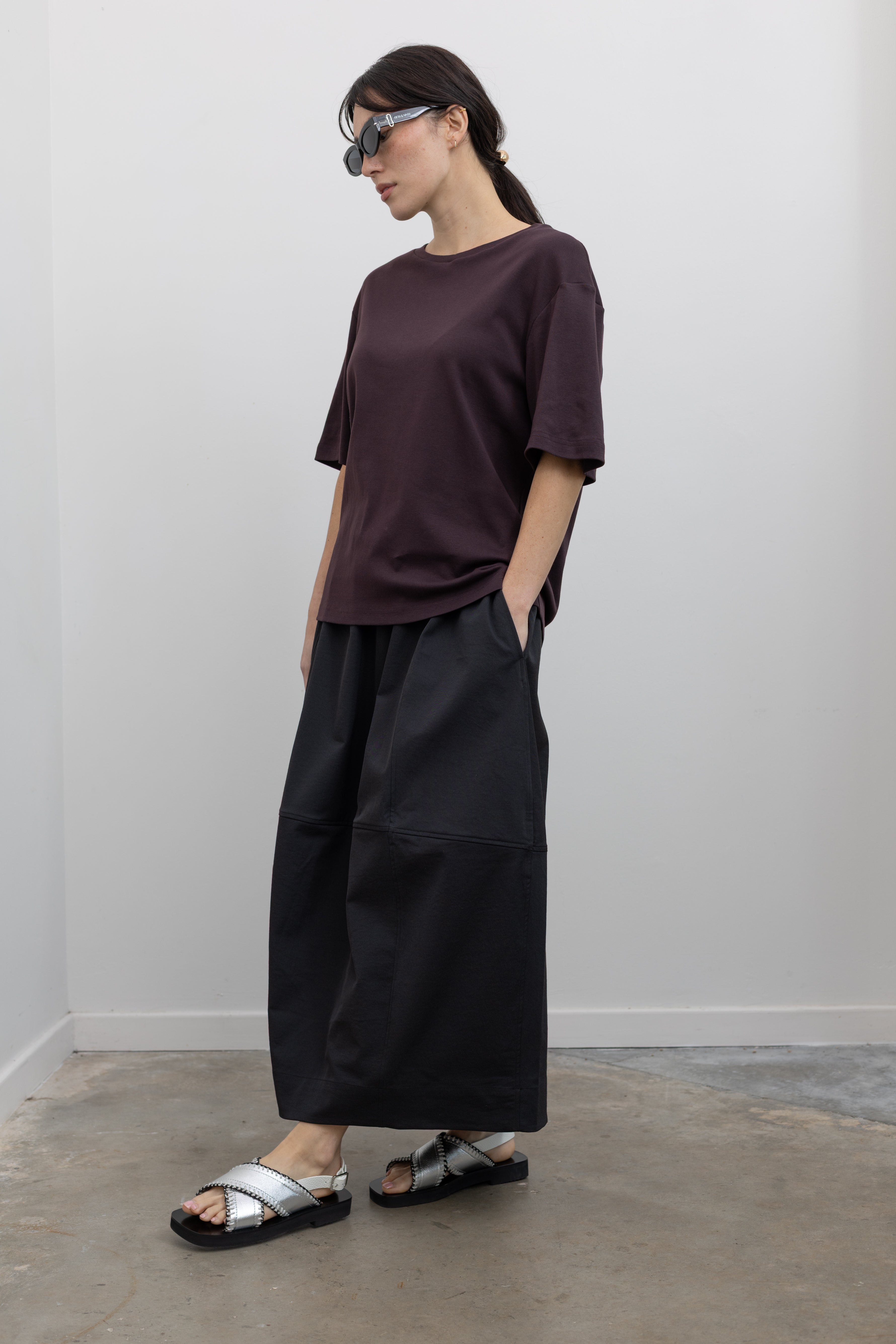 Full length 3/4 view of a woman standing wearing contemporary womenswear spring fashion in a Mijeong Park dark brown oversized t-shirt with a black voluminous balloon midi-to-maxi silhouette skirt featuring an elastic waistband paired with silver metallic slip-on sandals at an Alas Eius store. 