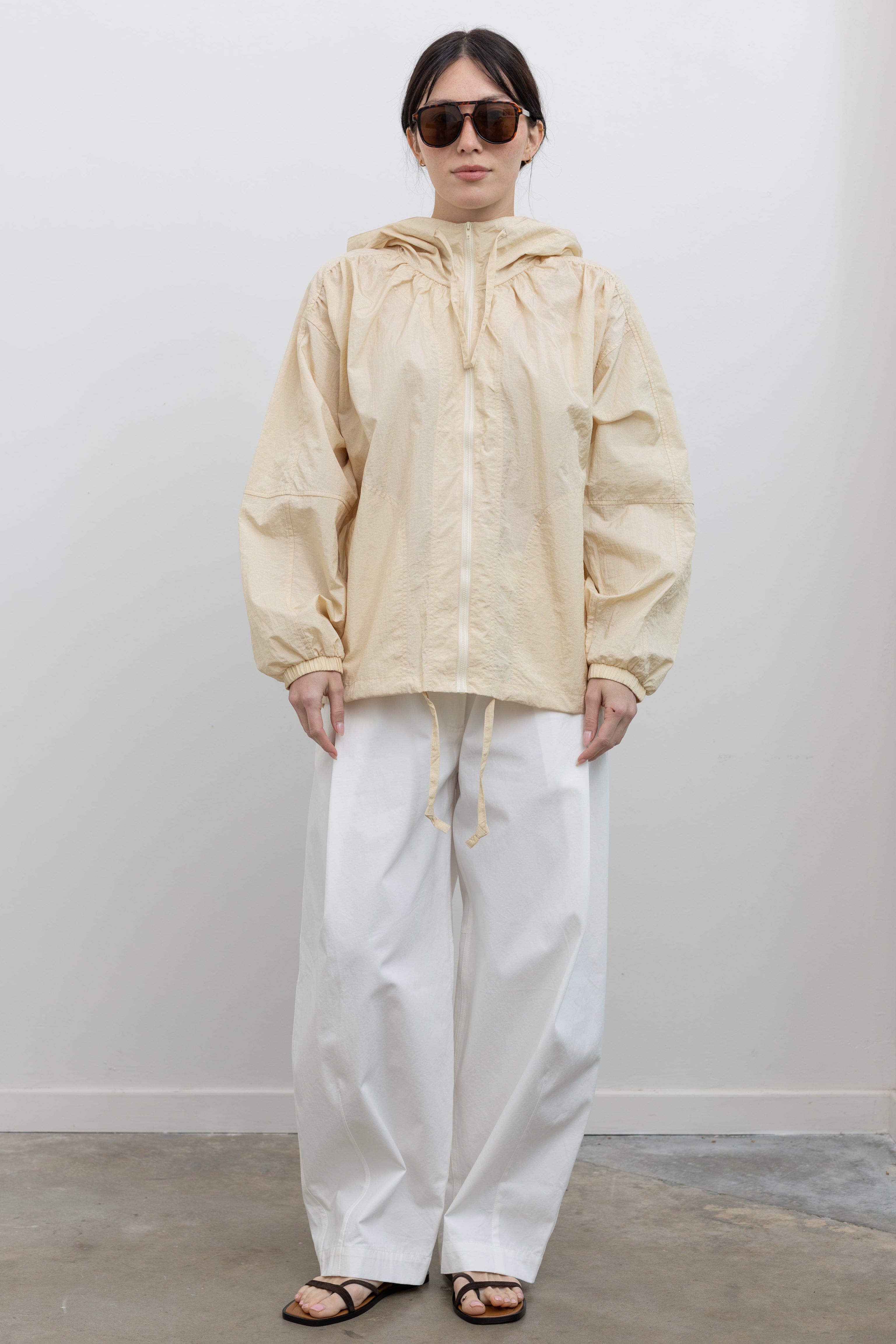 Front view of a woman posing with sunglasses wearing an elevated minimalist outerwear staple in a Mijeong Park cream colored nylon shirred anorak jacket featuring an oversized fit, front pockets, hood, and a zipper closure paired with white curved leg pants at an Alas Eius store. 