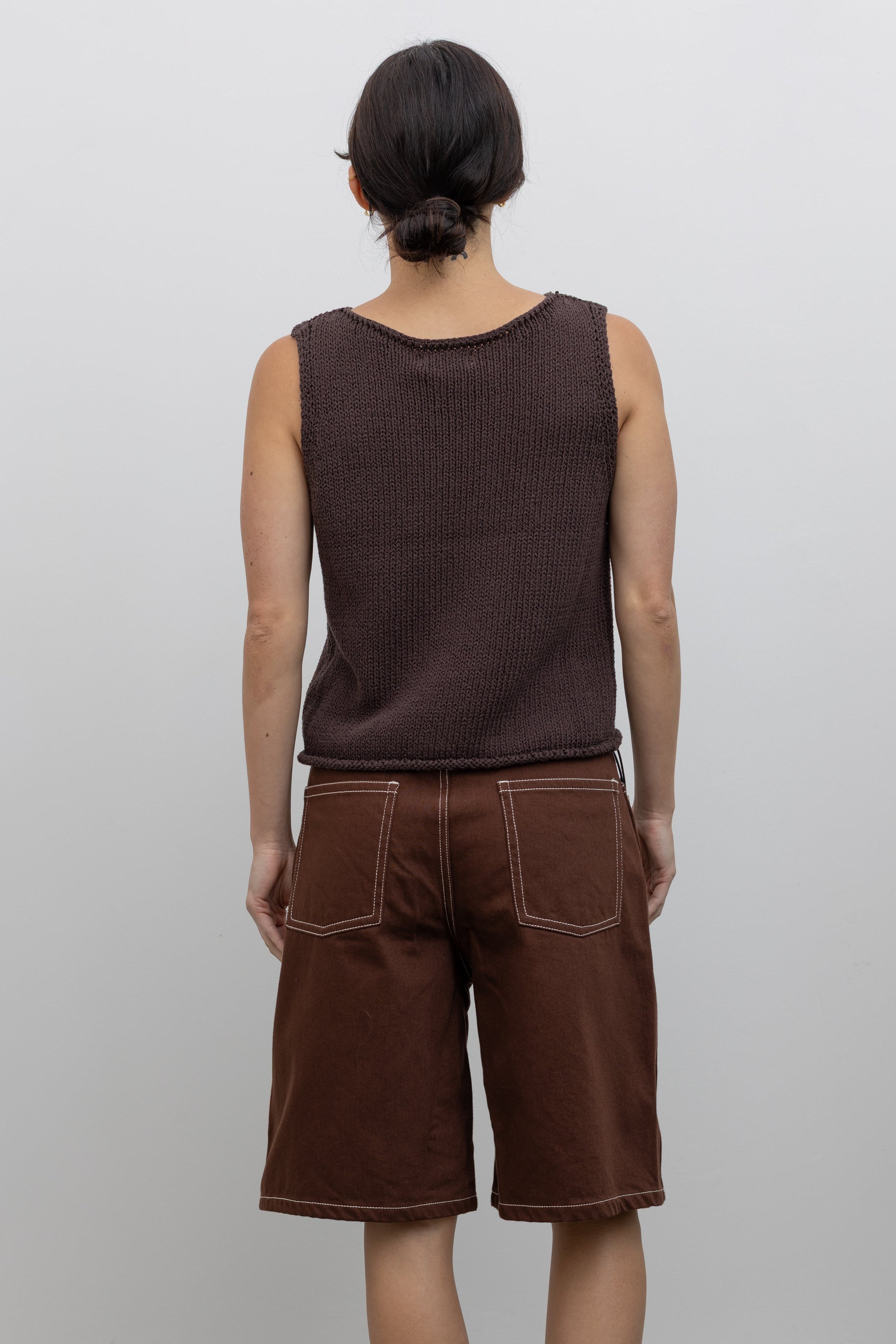 Full length back view of a woman posing wearing modern minimalist spring and summer fashion in a Mijeong Park dark brown boat neck knit tank featuring a relaxed fit and rolled edges at the neck and hem paired with Kowtow Framework Organic Cotton Brown Denim Long Shorts that sit just above the knee and feature white contrast stitching at an Alas Eius store.  