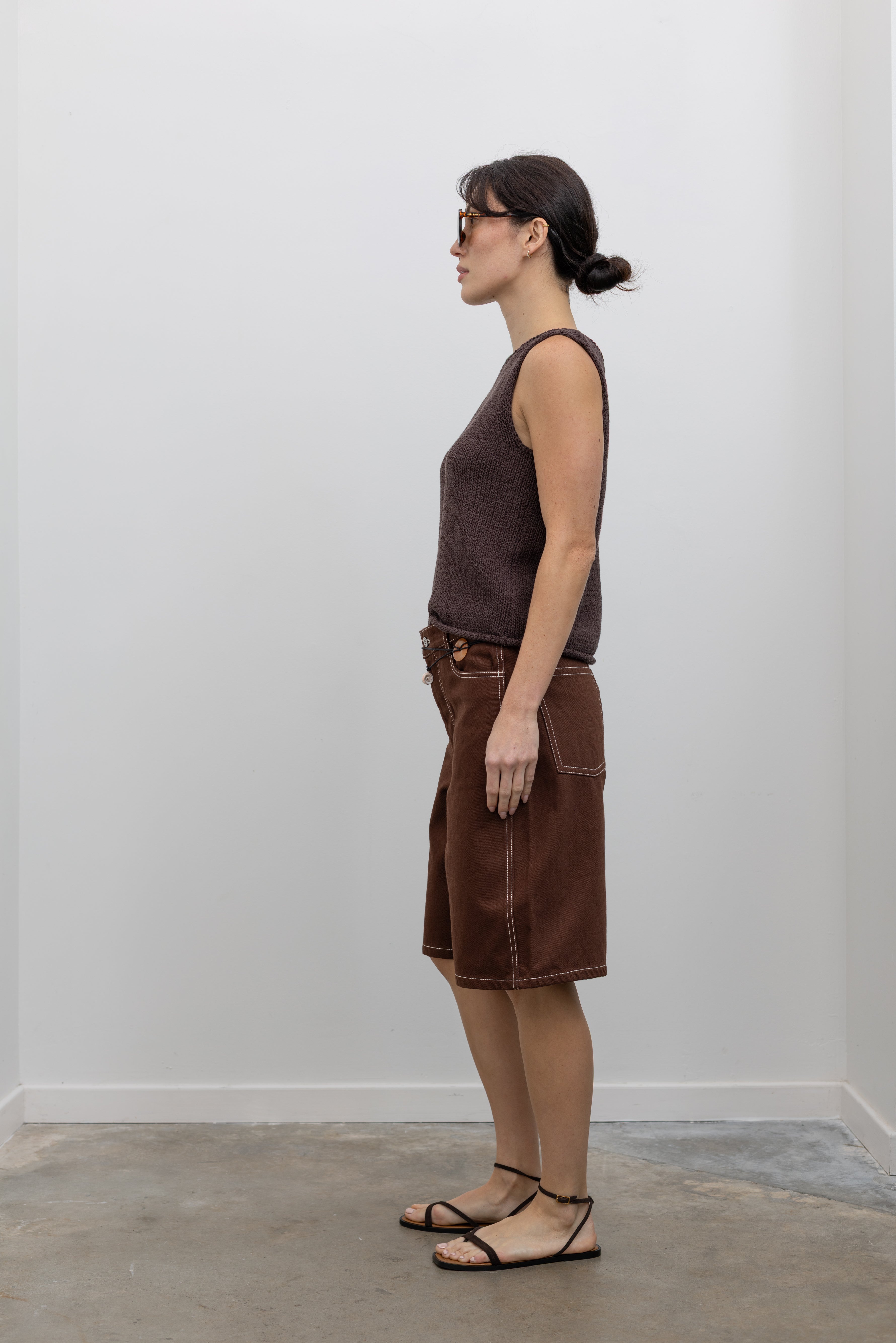 Full length side view of a woman posing wearing modern minimalist spring and summer fashion in a Mijeong Park dark brown boat neck knit tank featuring a relaxed fit and rolled edges at the neck and hem paired with Kowtow Framework Organic Cotton Brown Denim Long Shorts that sit just above the knee and feature white contrast stitching at an Alas Eius store.  
